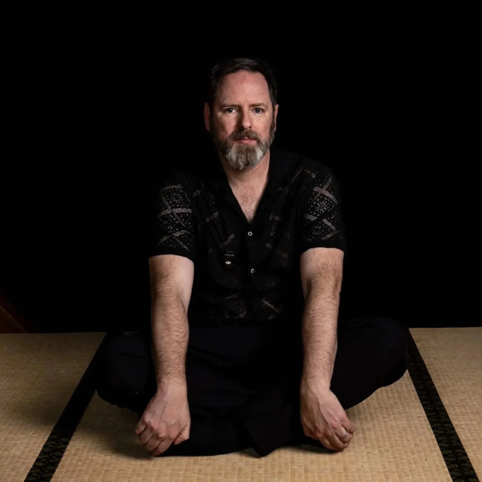 A man with a beard sitting cross-legged on a tatami mat floor, with his arms resting on his knees and hands relaxed. He is wearing a black patterned shirt and looks directly at the camera, set against a plain black background.