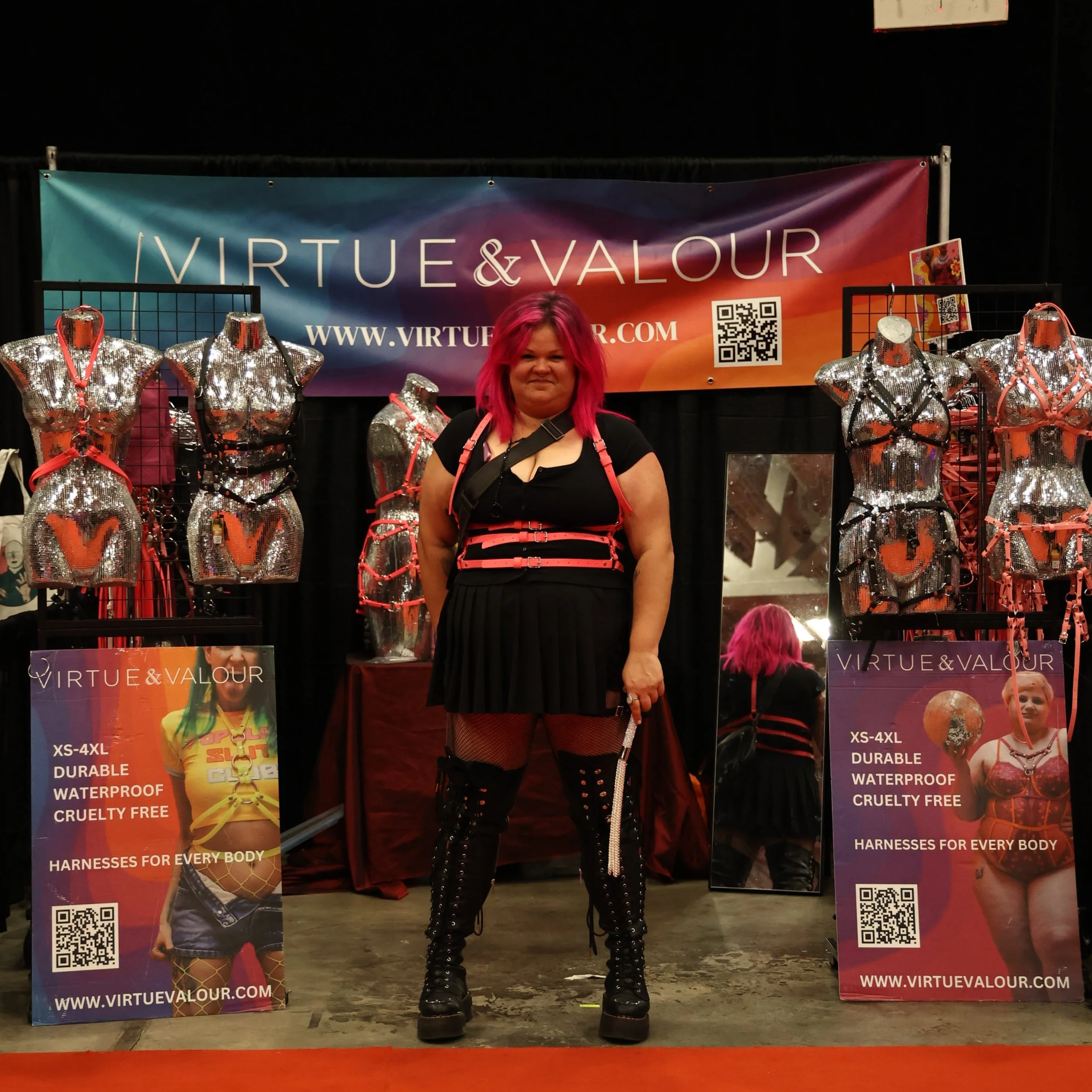 A woman with pink hair standing in front of a display booth with mannequins dressed in shiny, metallic, cut-out fashion outfits. The booth features a colorful banner with the text 'Virtue & Valour' and various promotional posters with QR codes and website URL virtuousalour.com.