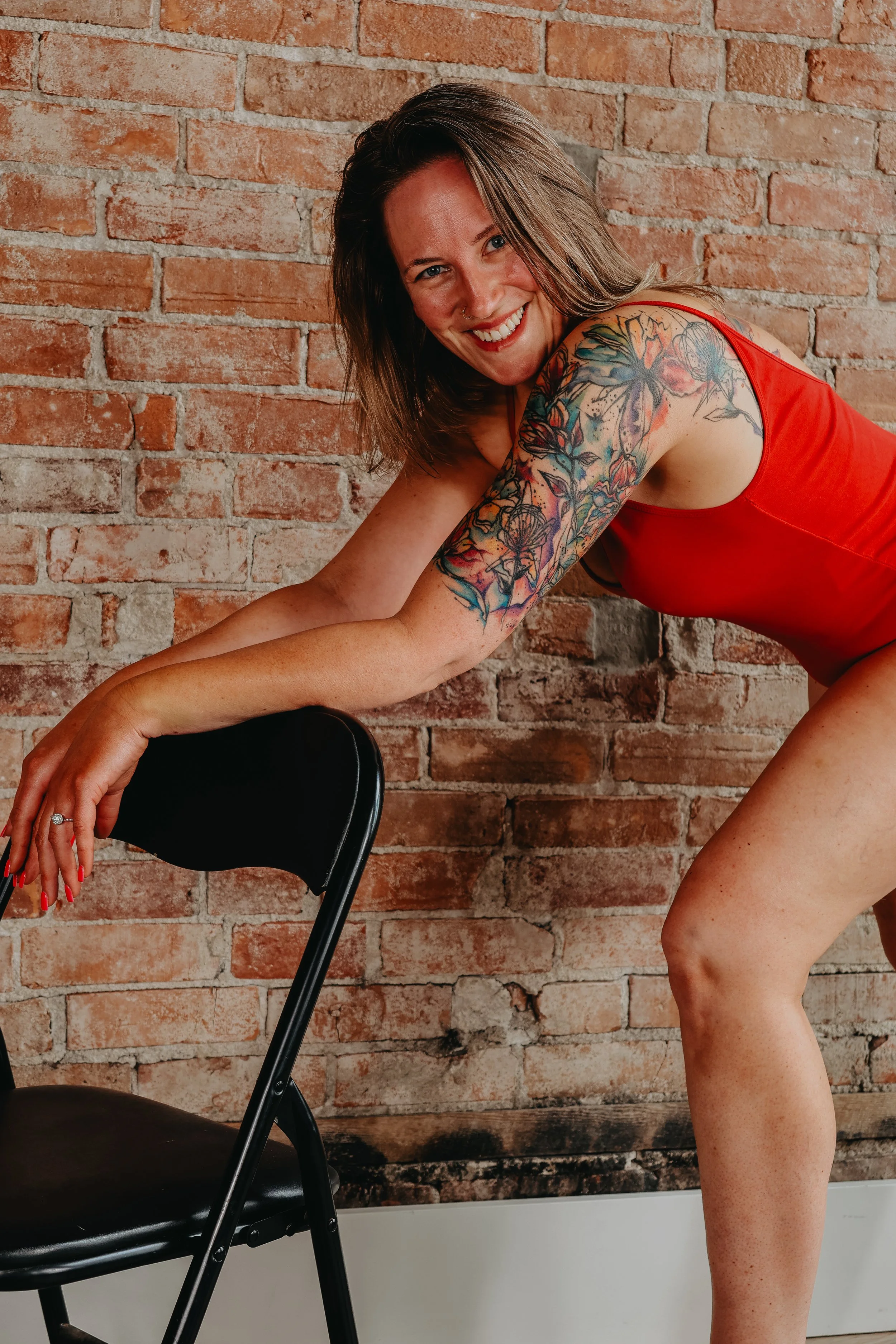 A smiling woman with shoulder-length brown hair, tattoos on her right arm, and wearing a red sleeveless top, leaning on a black chair with a brick wall background.