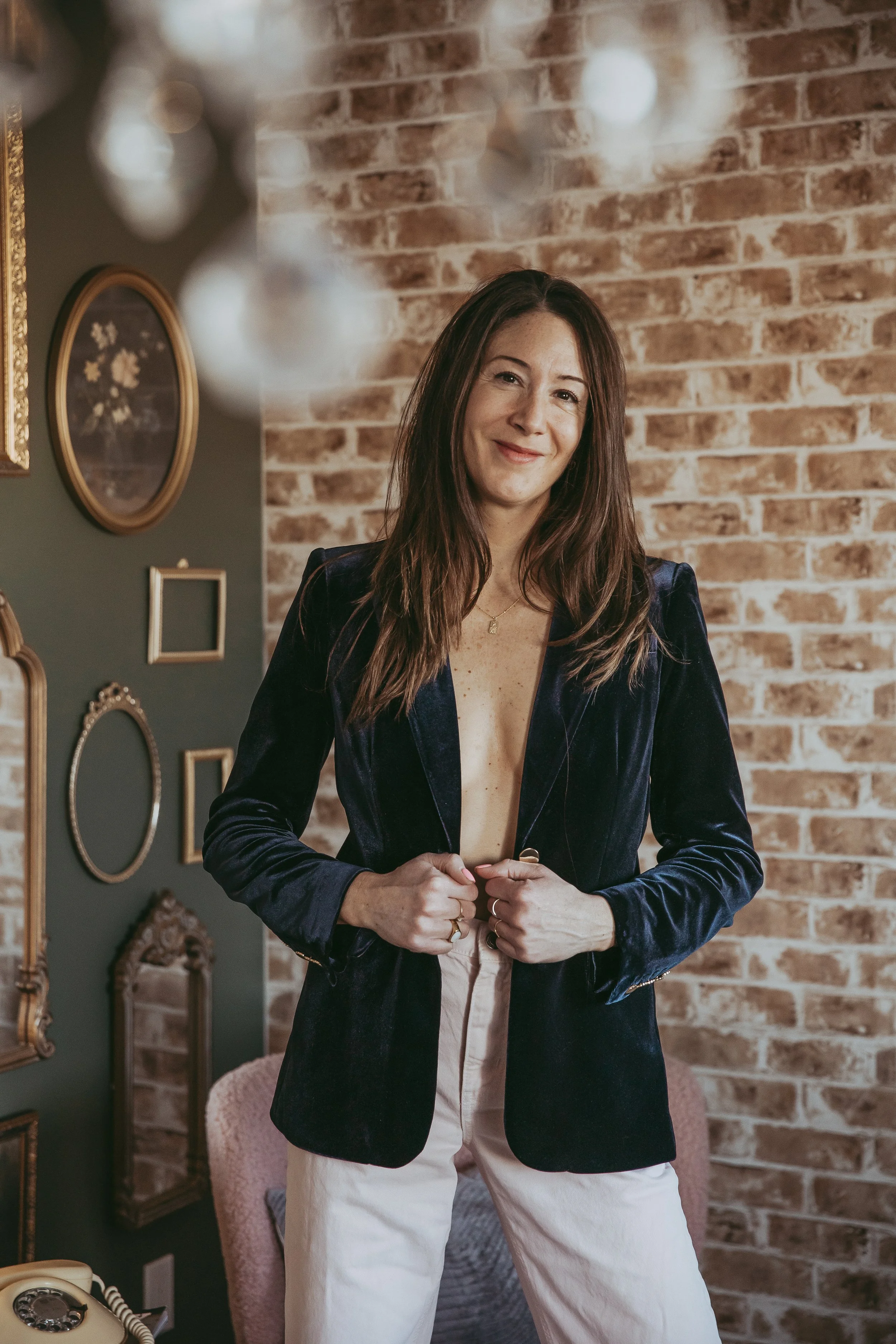 A woman standing in a room with a brick wall behind her, wearing a navy velvet blazer and light-colored pants, smiling at the camera.