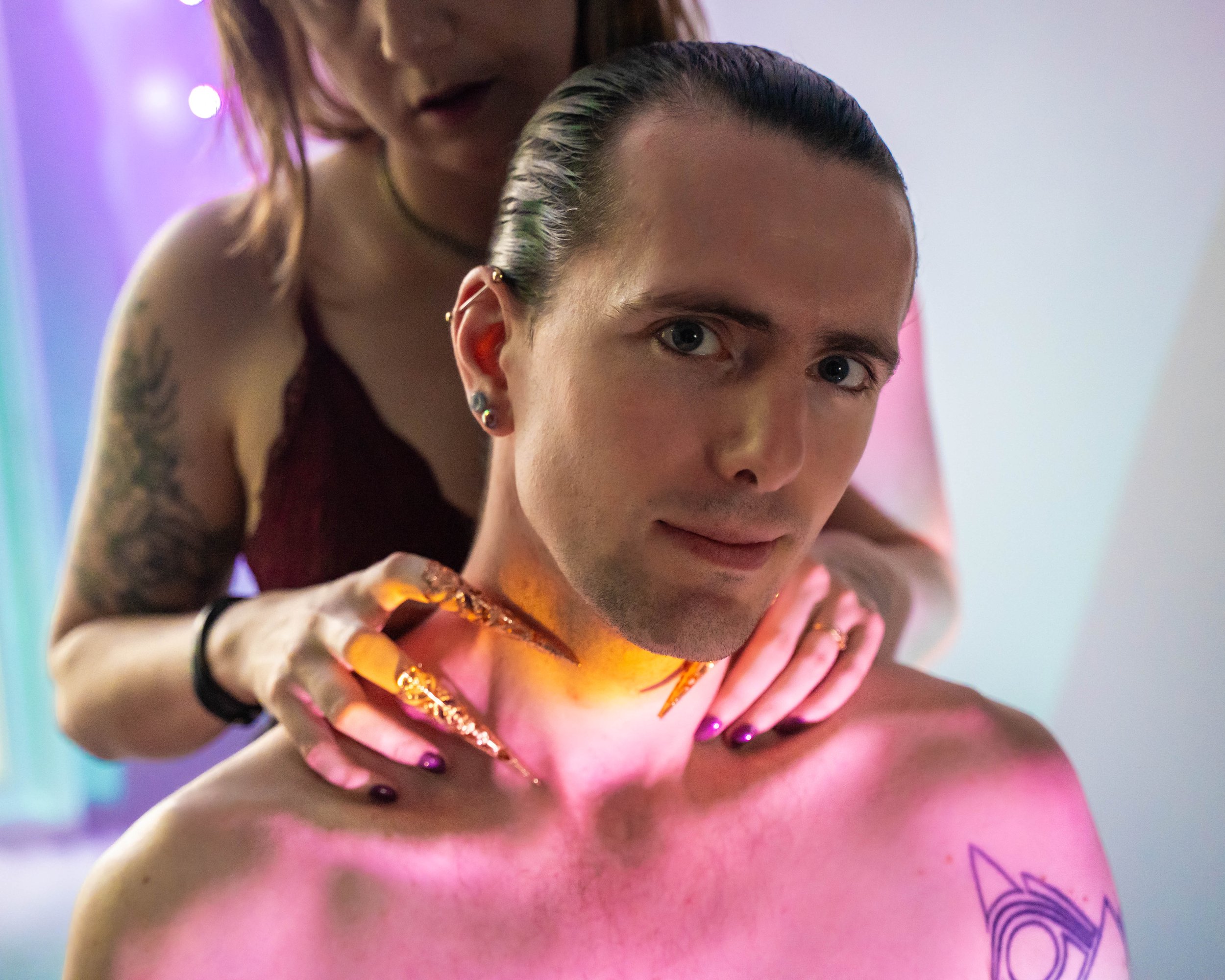 A woman with tattoos and earrings is leaning over a young person with short hair, who appears to be receiving a body tattoo or body painting with vibrant lighting effects.