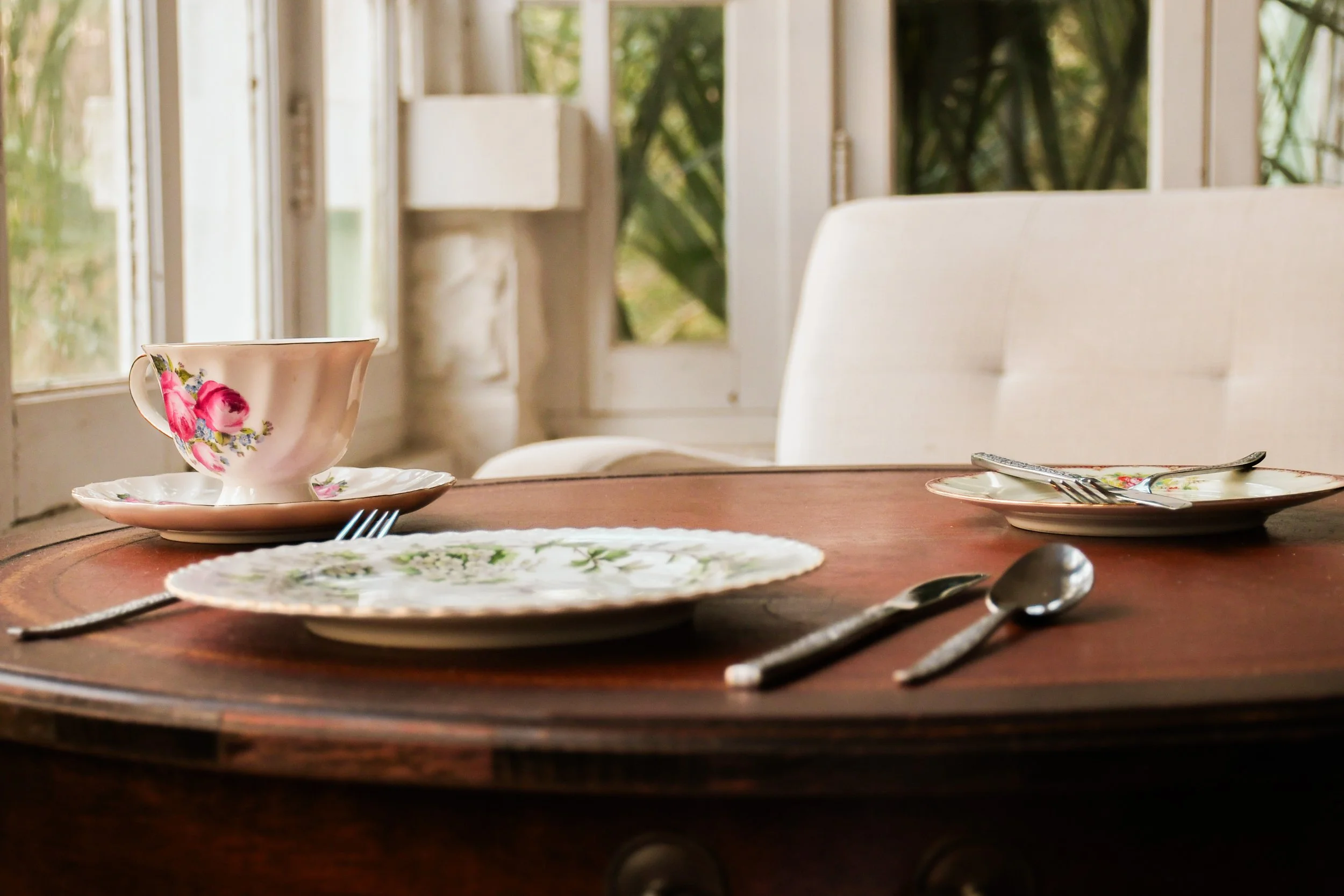 Dining table set with floral dishes, a teacup, a plate, a fork, a spoon, and a knife, with a white cushioned chair and windows in the background.