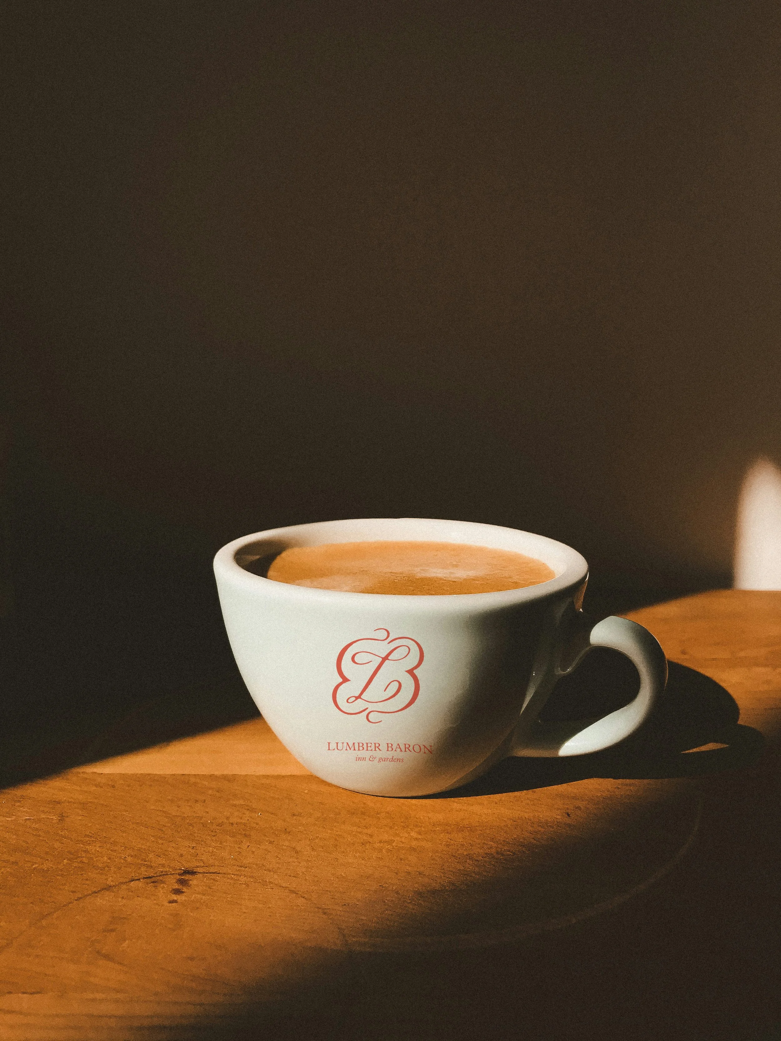 A white coffee cup with a red logo and text, filled with coffee, on a wooden surface with sunlight shining on it.