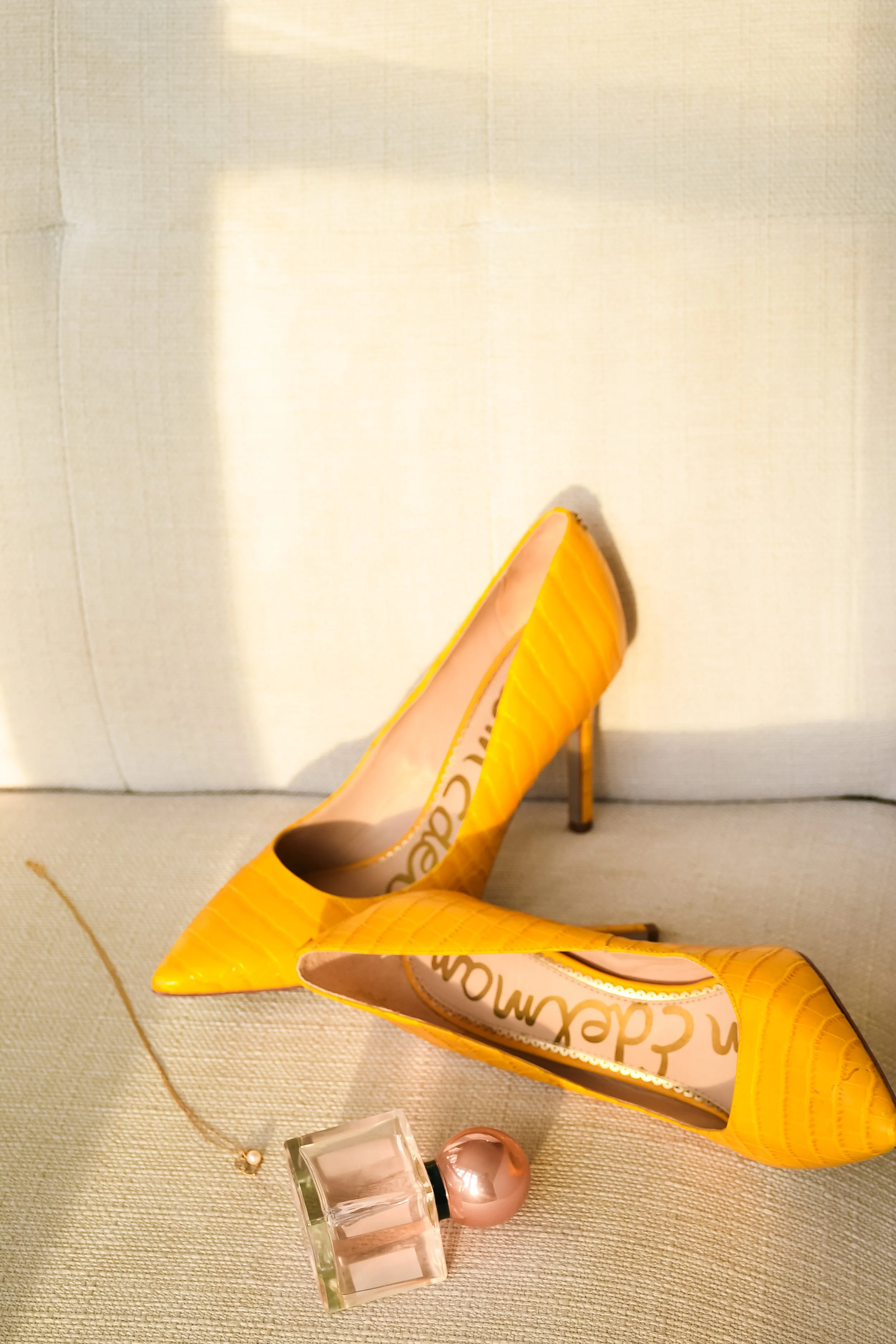 Yellow high-heeled shoes, a perfume bottle with a pink round cap, and a necklace with a pearl pendant on a beige surface.