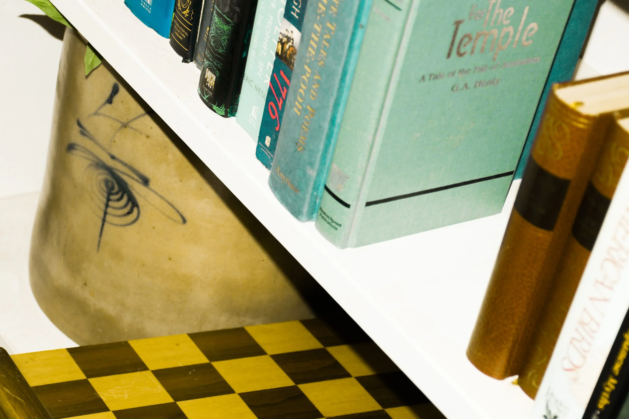 Close-up of a bookshelf displaying various books, with a decorative ceramic container featuring a face and glasses, and part of a wooden chessboard visible in the foreground.