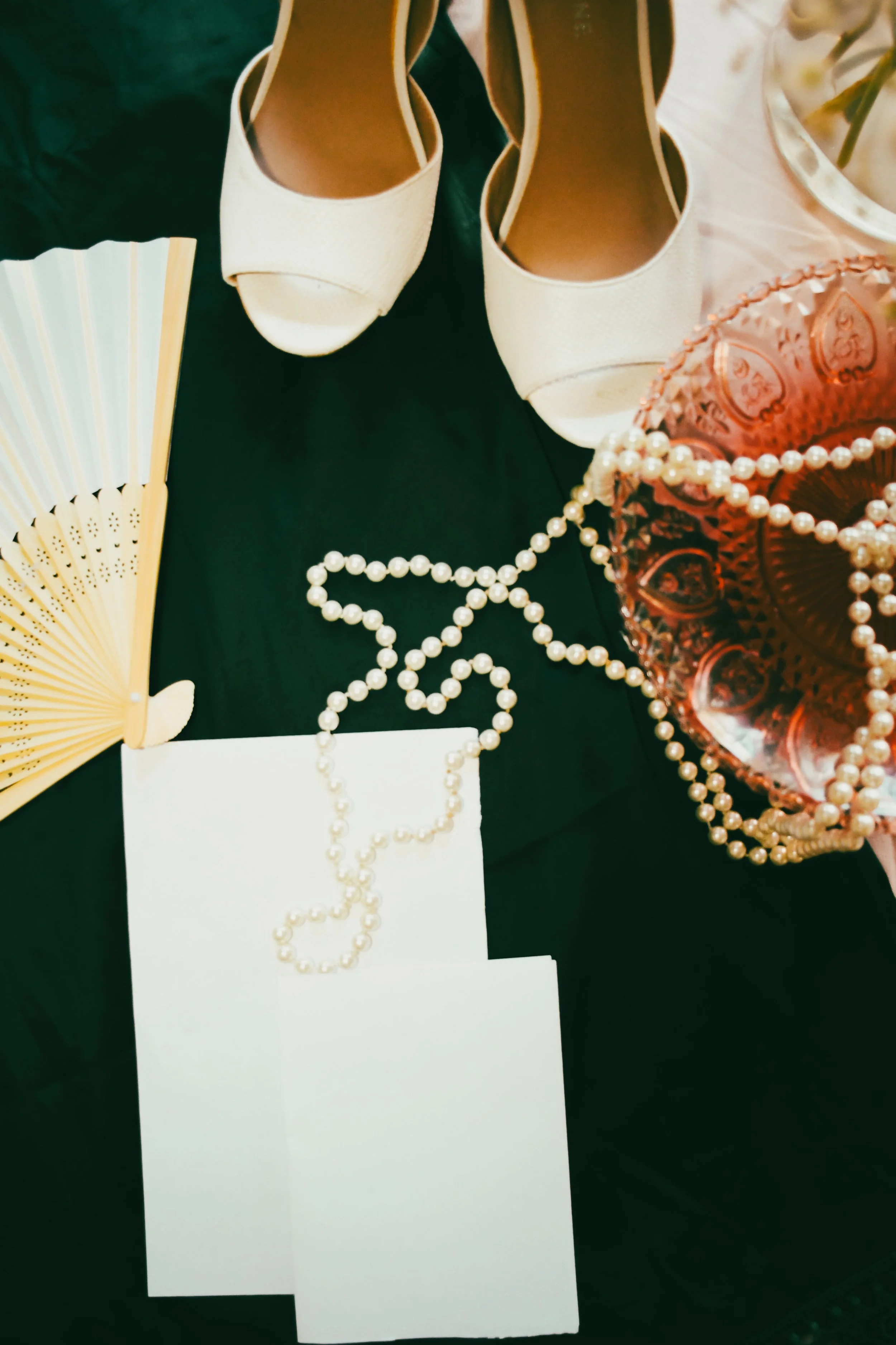 Pair of white high-heeled shoes, pink glass with pearl necklaces, a yellow fan, and blank white cards on a black surface.