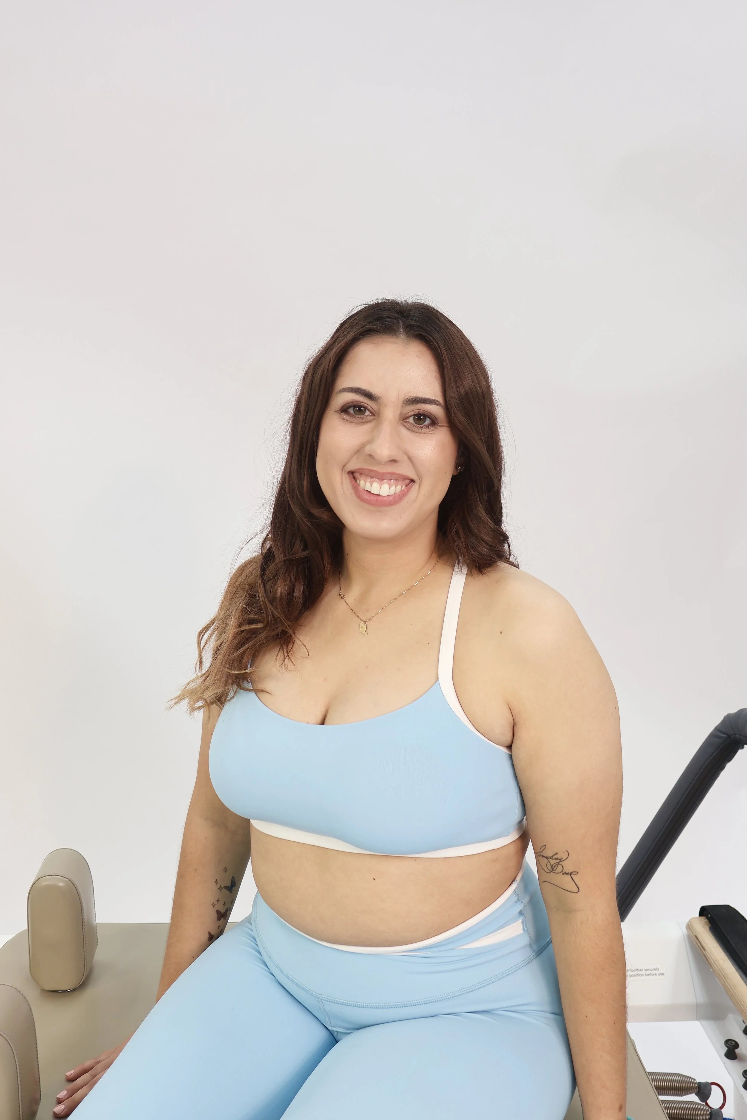 A woman with long brown hair, wearing a light blue sports bra and matching leggings, sitting on a beige medical examination table, smiling at the camera.