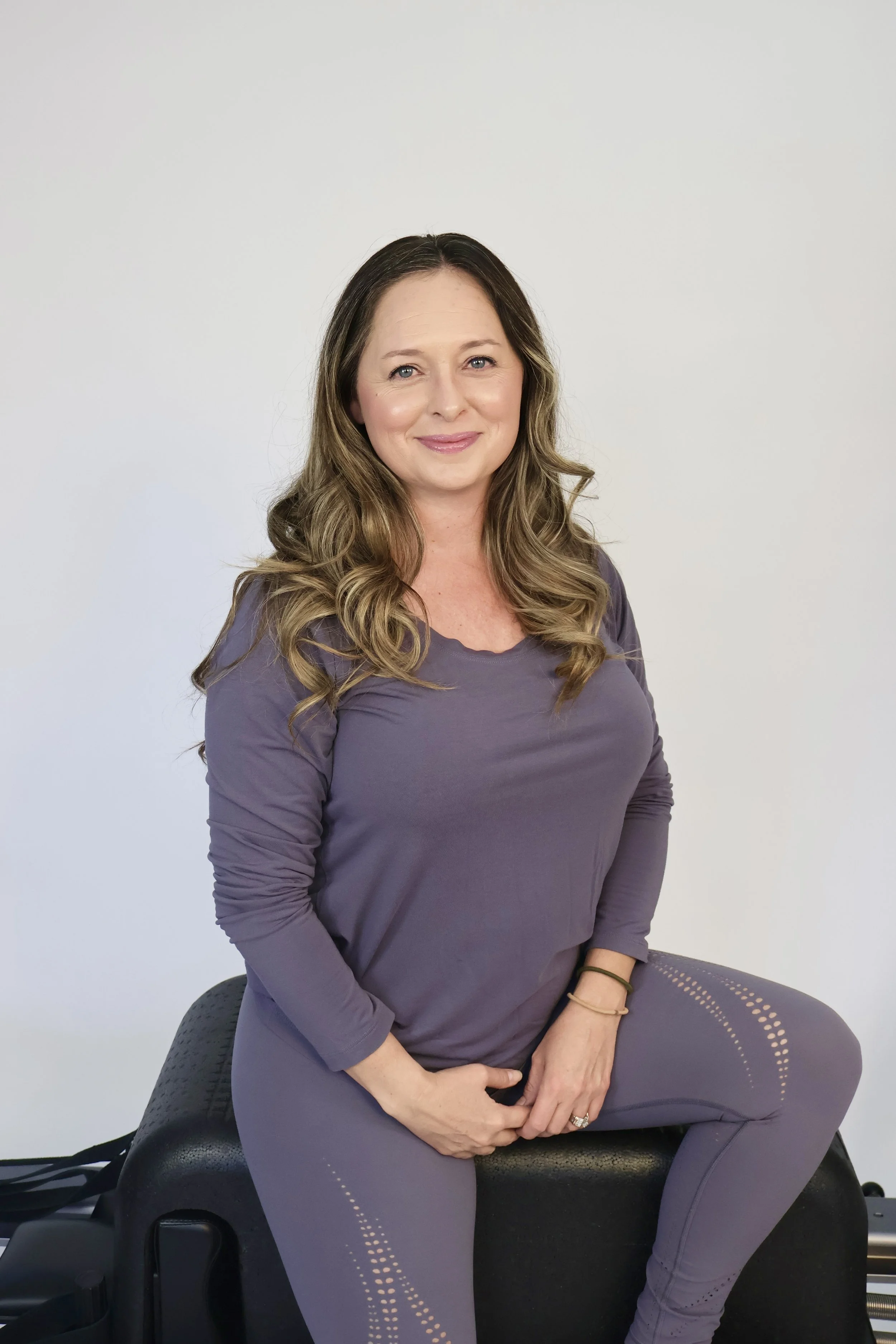 A woman with long wavy brown hair, smiling, wearing a purple workout outfit, sitting on exercise equipment against a plain white wall.