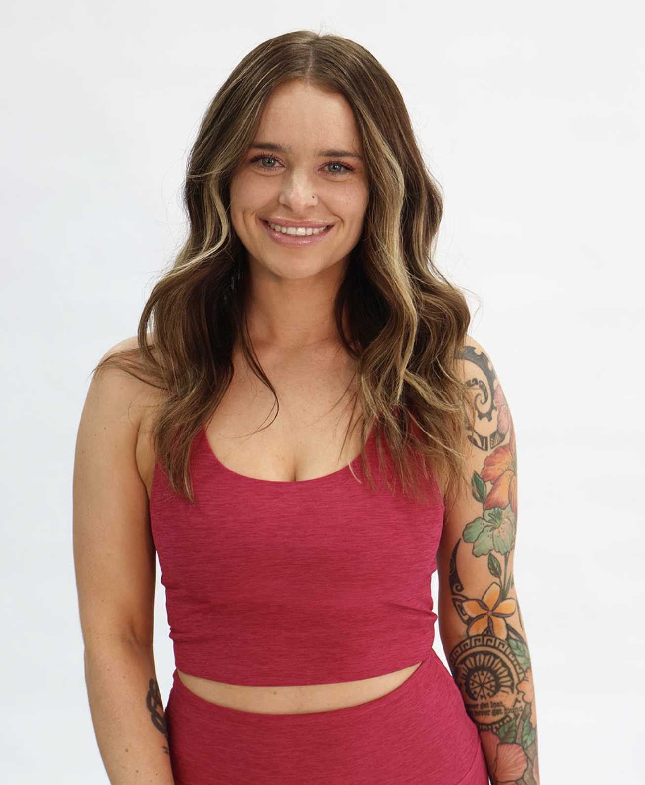 A woman with dark brown wavy hair smiling, wearing a red sports bra and matching leggings, kneeling on a Pilates reformer machine, with a white background.