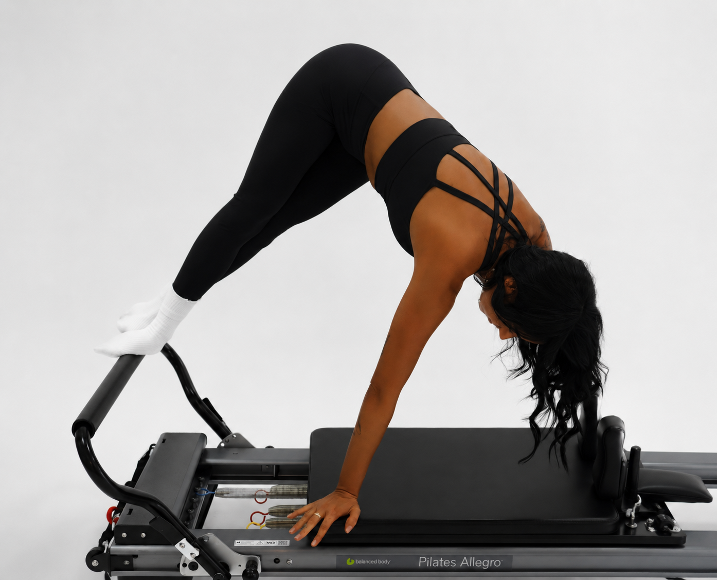 A woman in black sportswear performs a plank exercise on a Pilates reformer machine with her feet on the pulley side and hands on the platform.