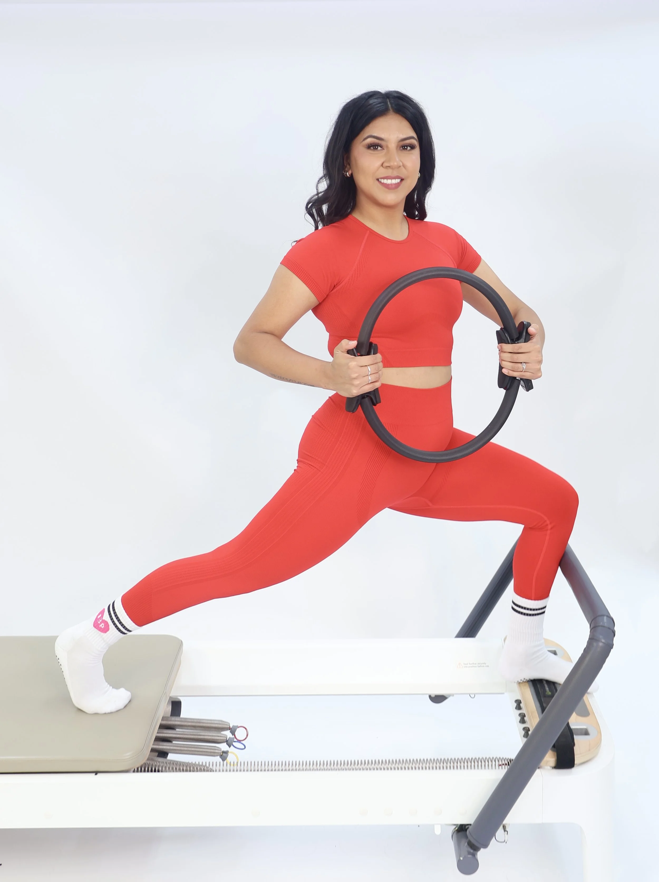 A woman in red workout clothes stretching on a Pilates reformer machine, holding a Pilates ring.