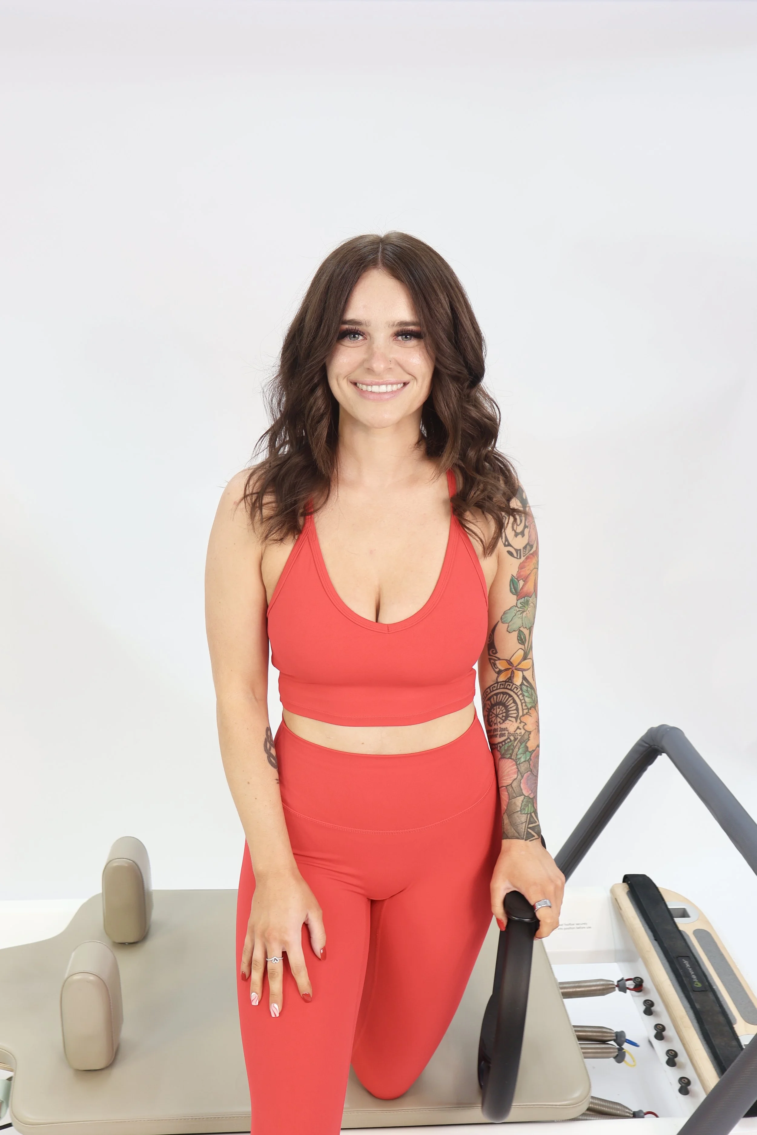 A woman with dark brown wavy hair smiling, wearing a red sports bra and matching leggings, kneeling on a Pilates reformer machine, with a white background.
