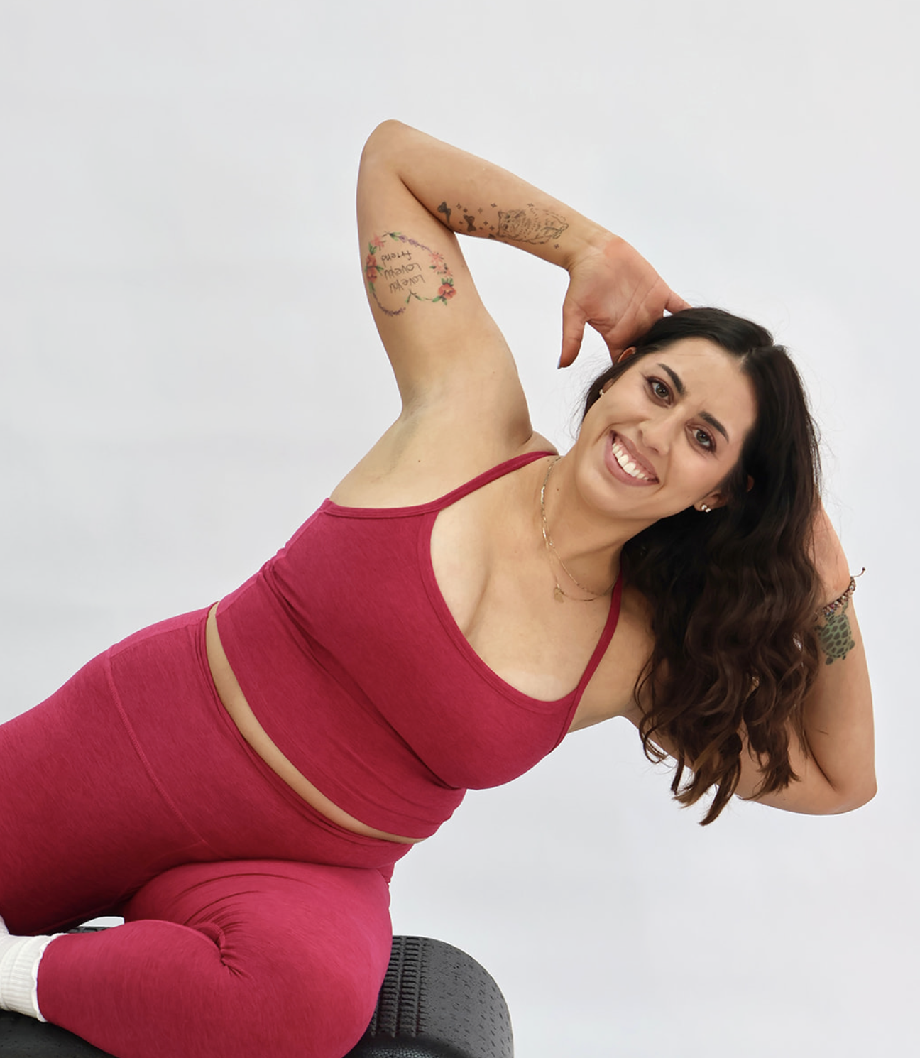 A woman with long brown hair, wearing a light blue sports bra and matching leggings, sitting on a beige medical examination table, smiling at the camera.