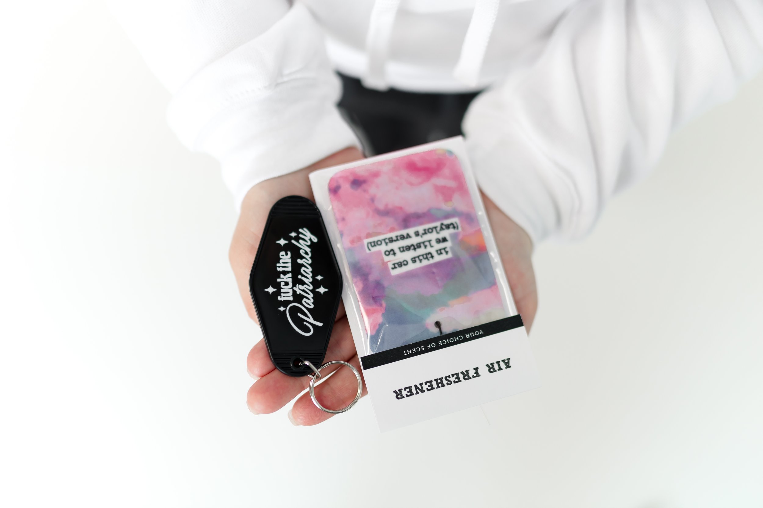 Person holding a 'Fuck the Patriarchy' keychain and a packaged air freshener with a colorful design.