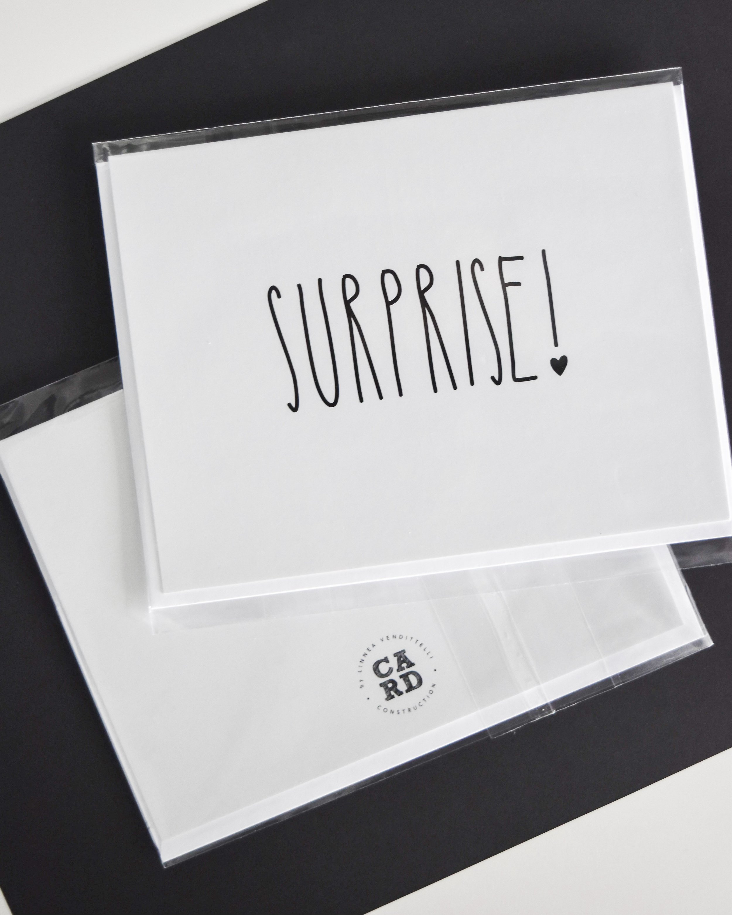 Greeting card with 'Surprise!' text and a small heart symbol, placed on a black background.