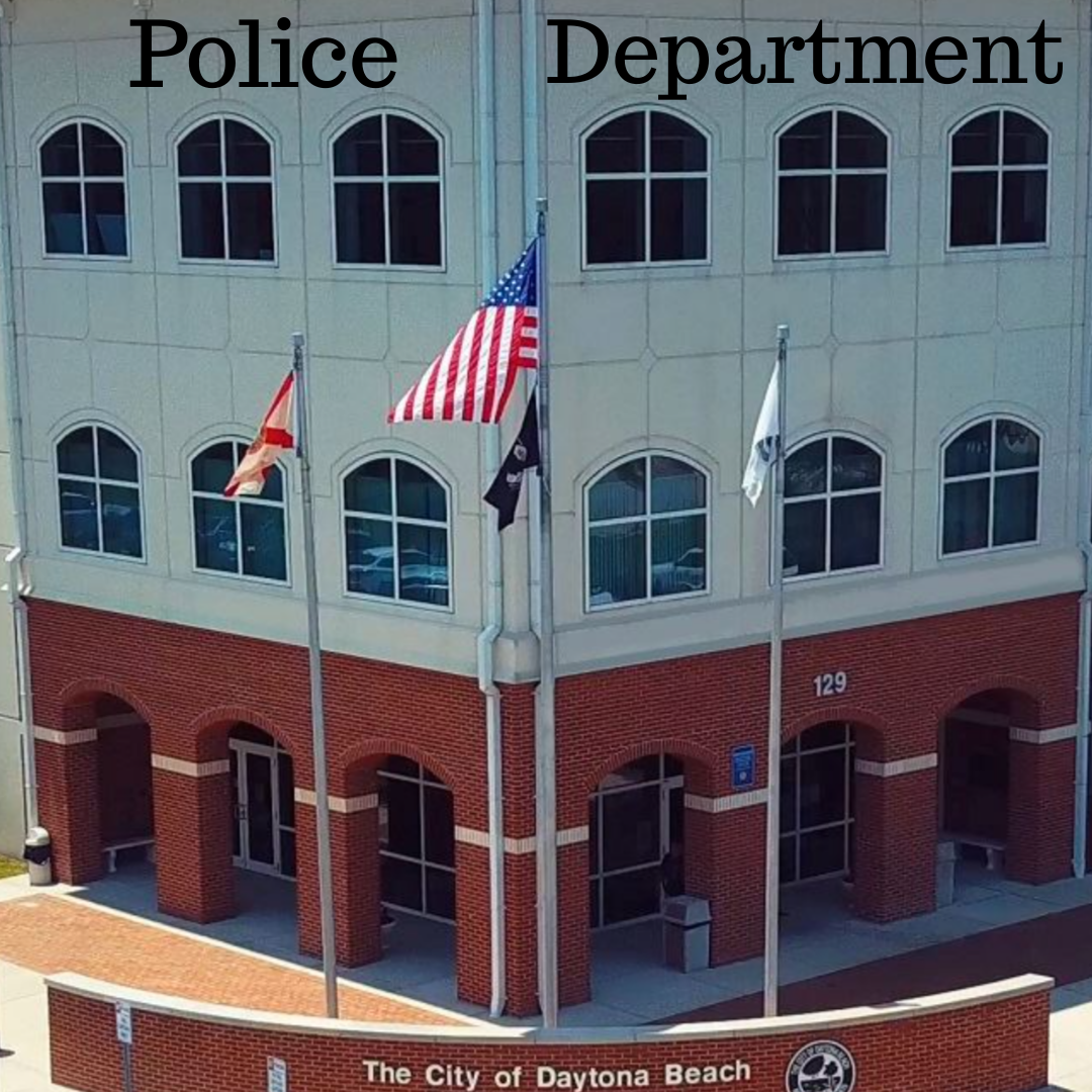 Front view of the Daytona Beach police department building with three flagpoles, the American flag, and two other flags, with a red brick base and white upper floors and windows, with the text "The City of Daytona Beach" at the bottom.