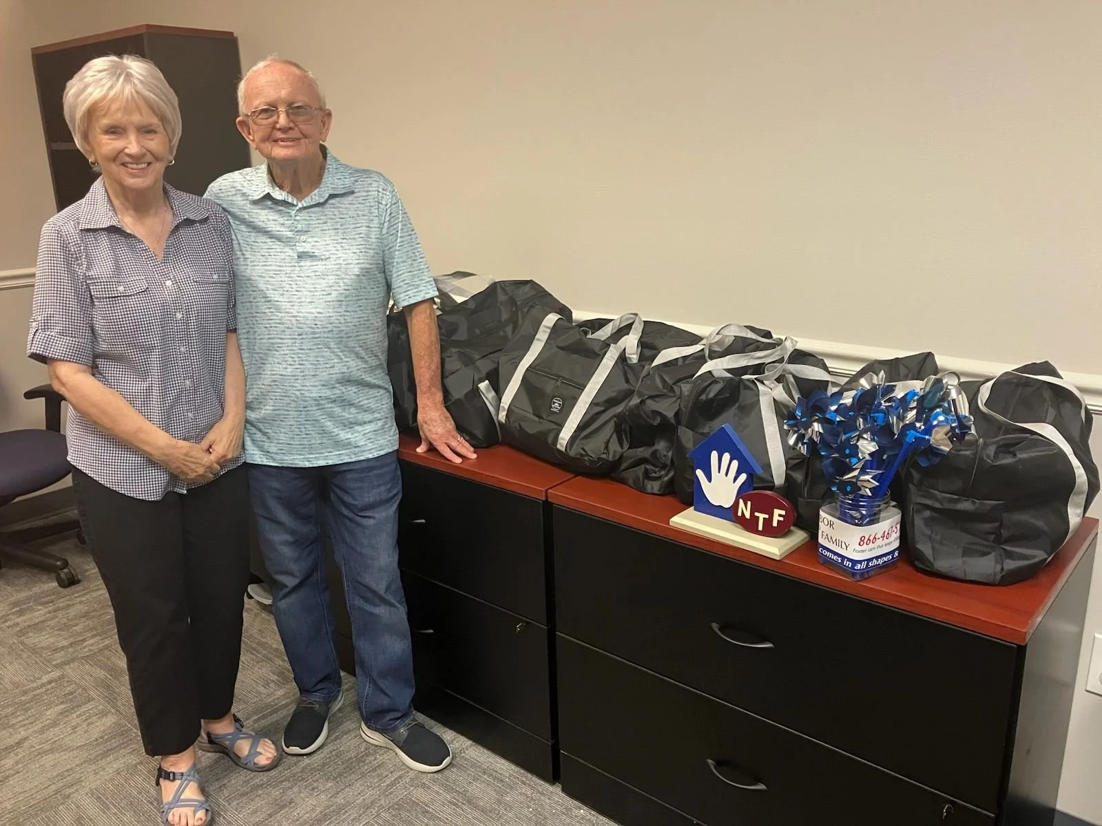 Two elderly people small woman and man smiling and standing to the side of a desk with several large black bags, a cup with pens, and decorative items on top of the desk.