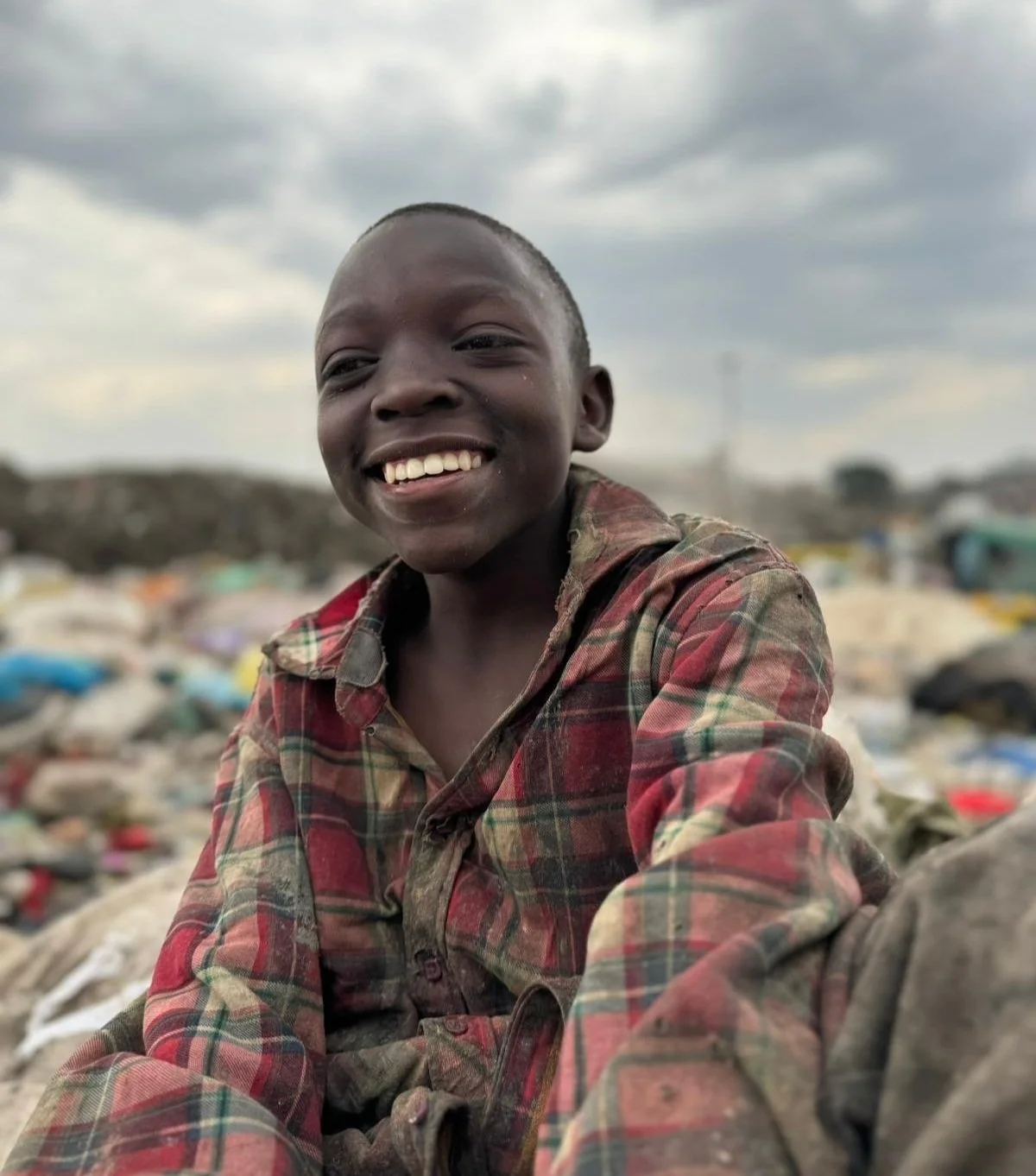 A smiling child in a red plaid shirt sits outdoors with a cloudy sky in the Dandora Landfill in Kenya.