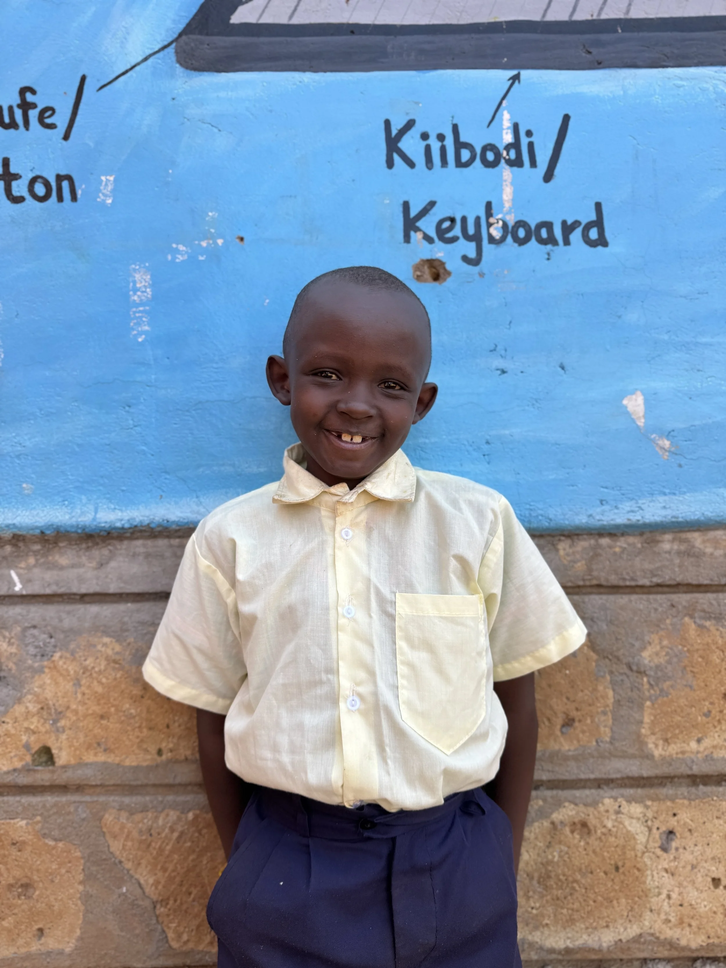 Smiling child in a light colored shirt standing against a blue wall with text "Kiibodi / Keyboard."