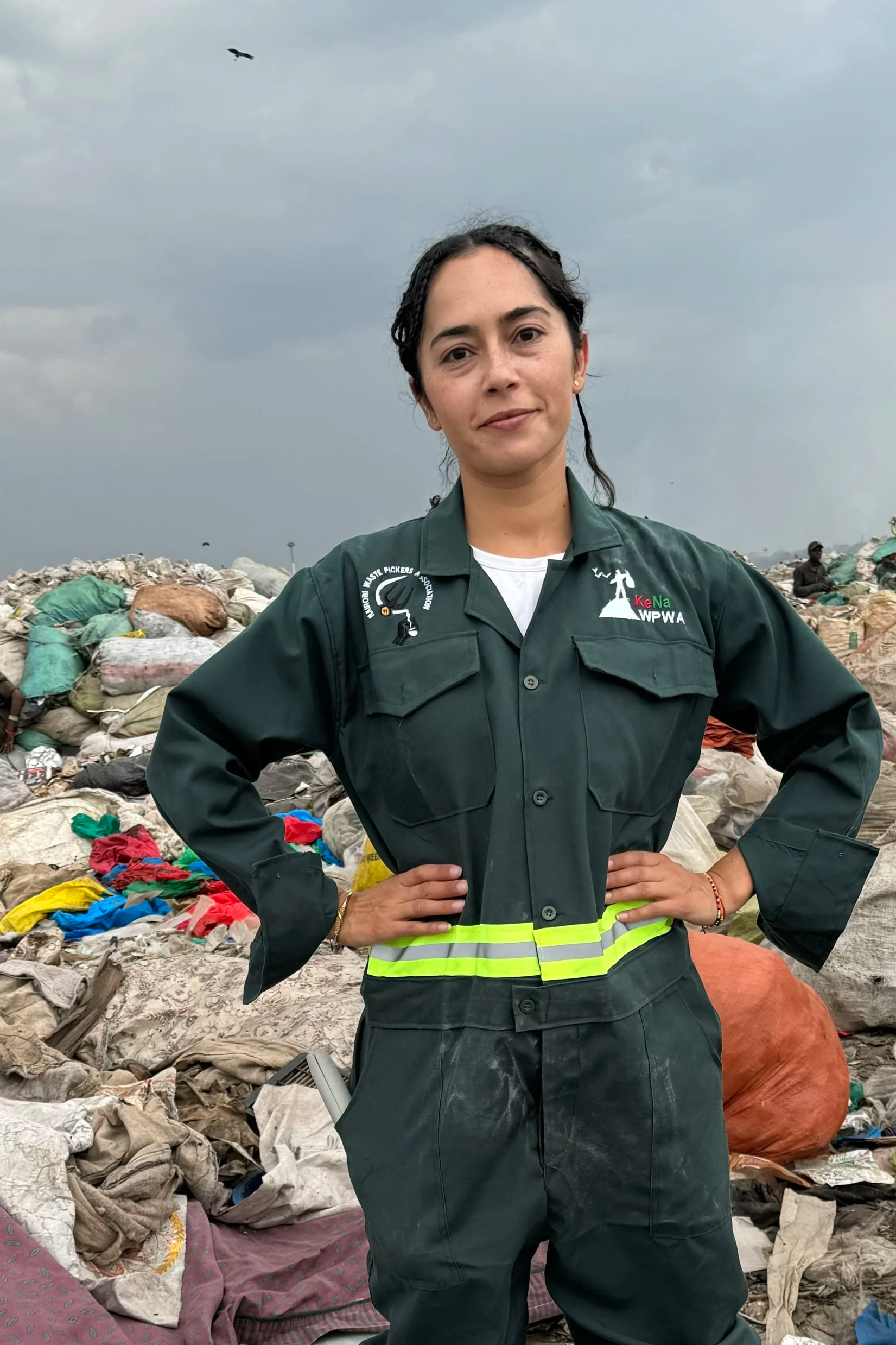 A woman in a uniform standing with hands on hips in a landfill filled with trash and debris, under cloudy skies.