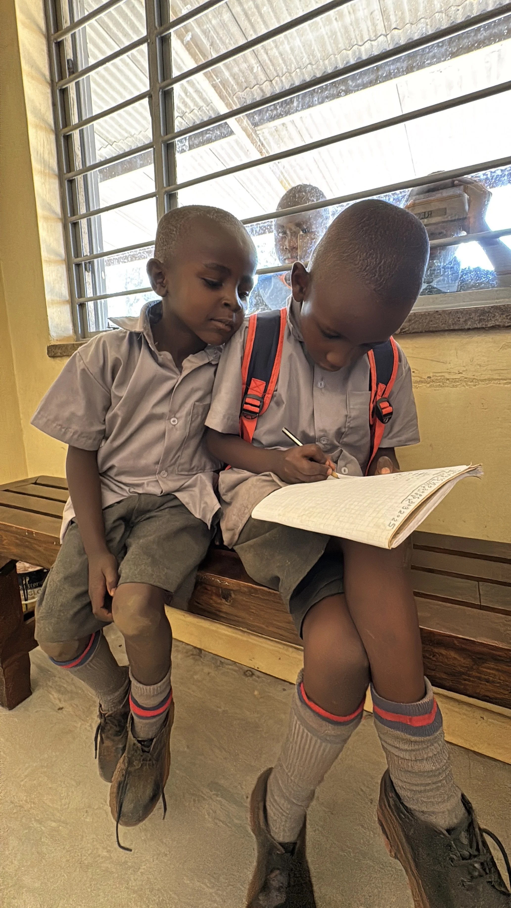 Two schoolchildren sitting on a bench, one writing in a notebook. Both wear gray uniforms and backpacks.