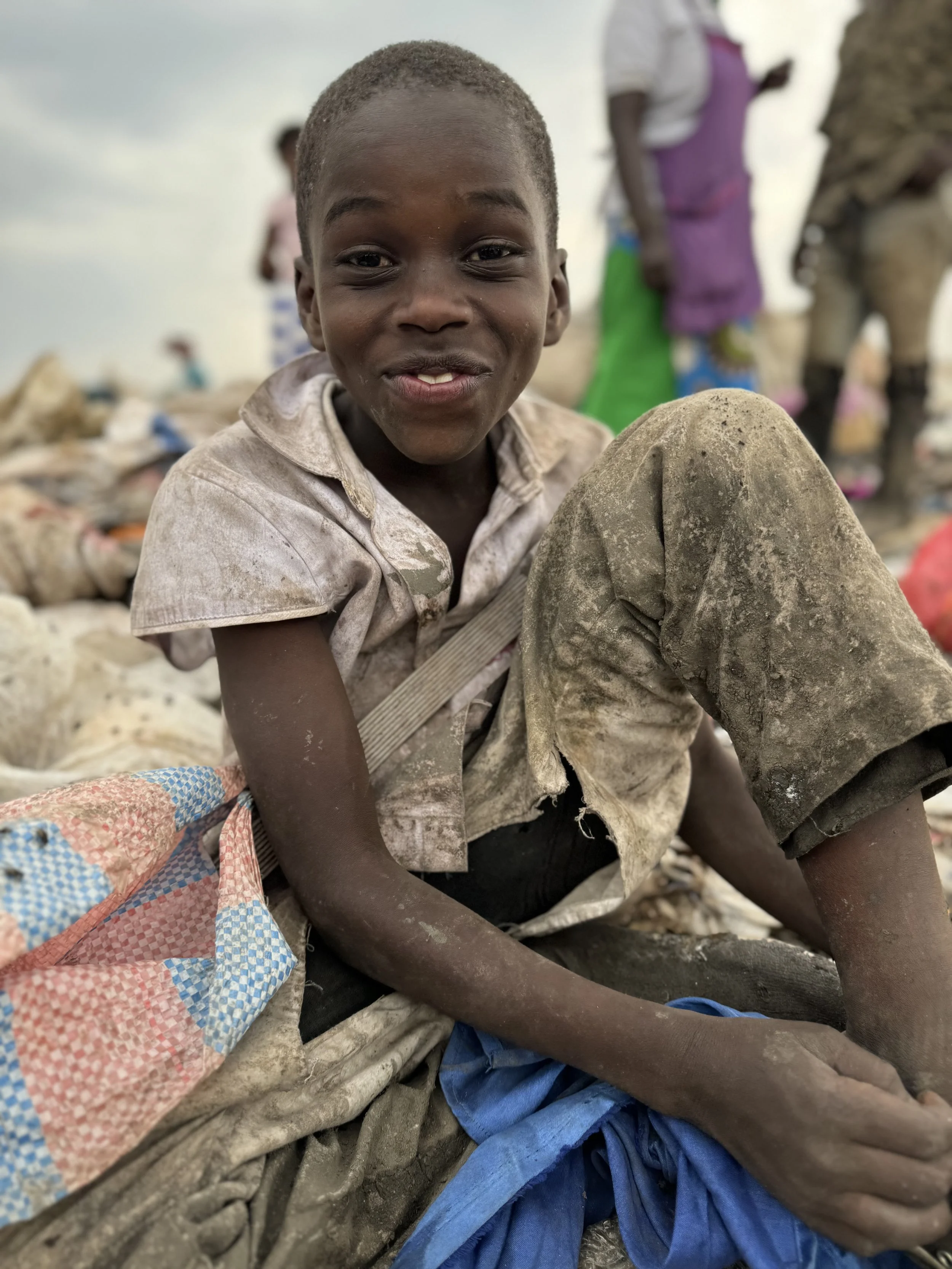 Smiling child in worn clothes sitting outdoors, with colorful fabric around them in the Dandora Landfill in Kenya.