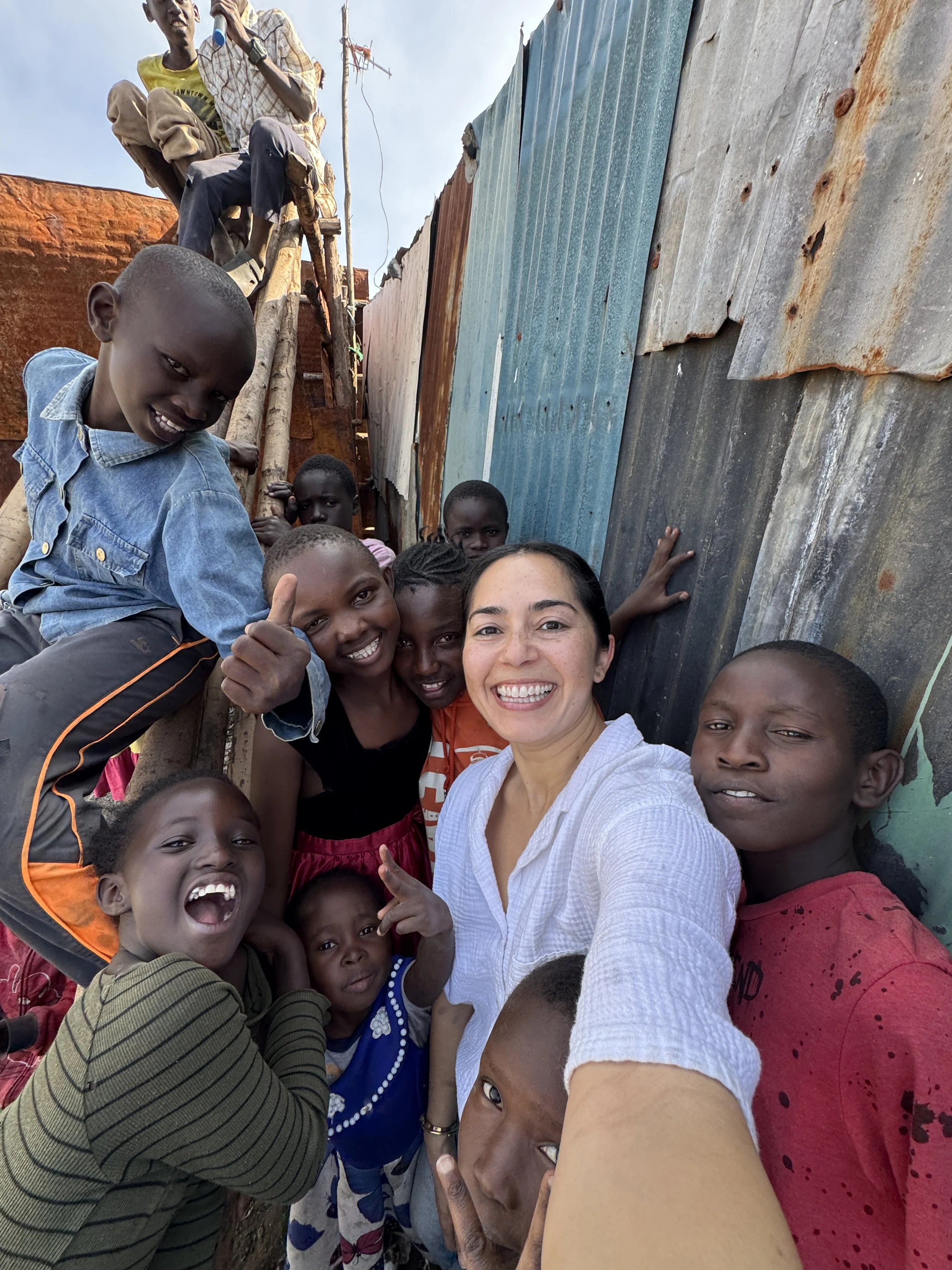 A group of smiling children and a woman taking a selfie together in a community setting with makeshift walls and a ladder in the background.