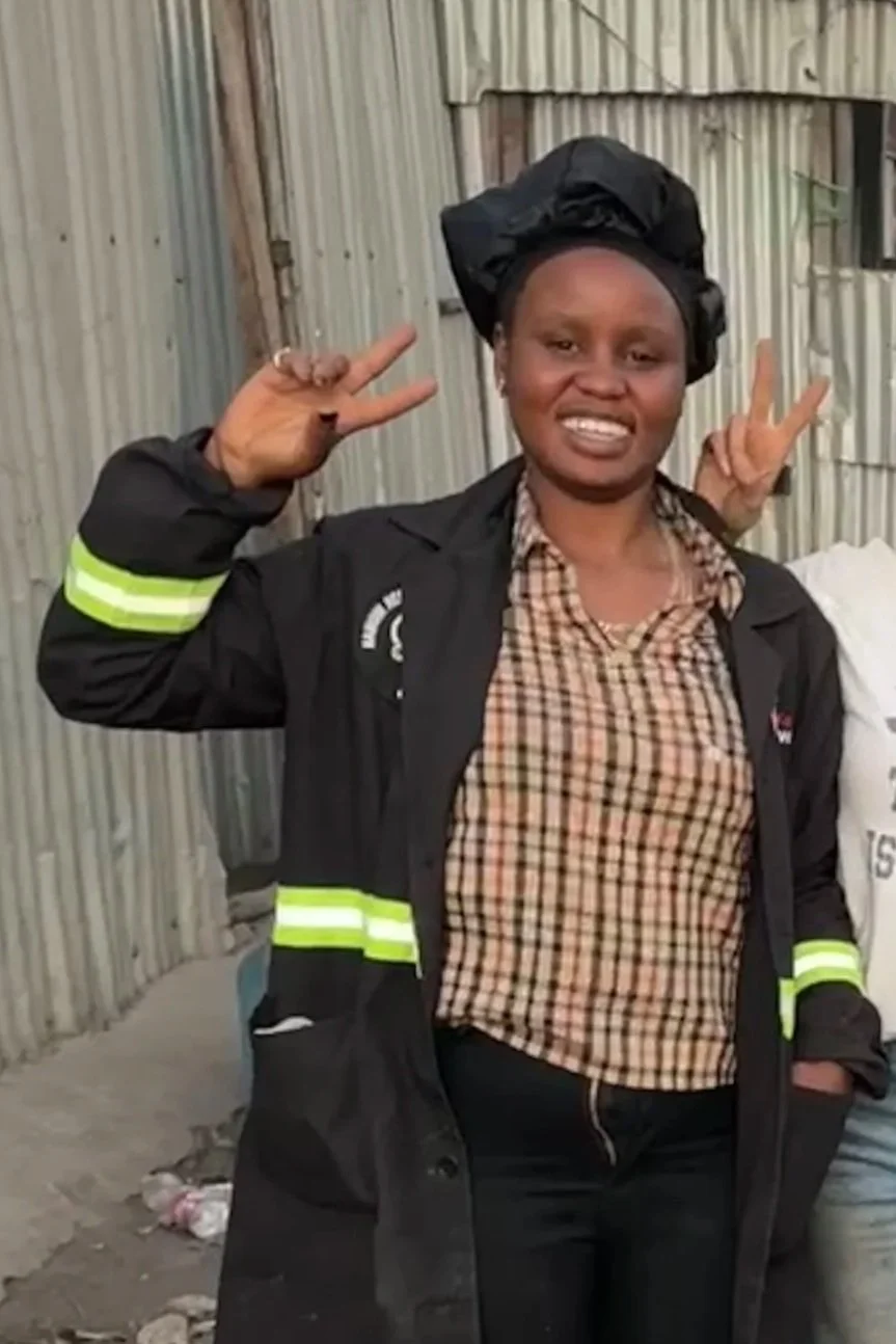 A woman smiling and making peace signs with both hands, wearing a black jacket with reflective stripes over a checked shirt, standing in front of a corrugated metal shed.