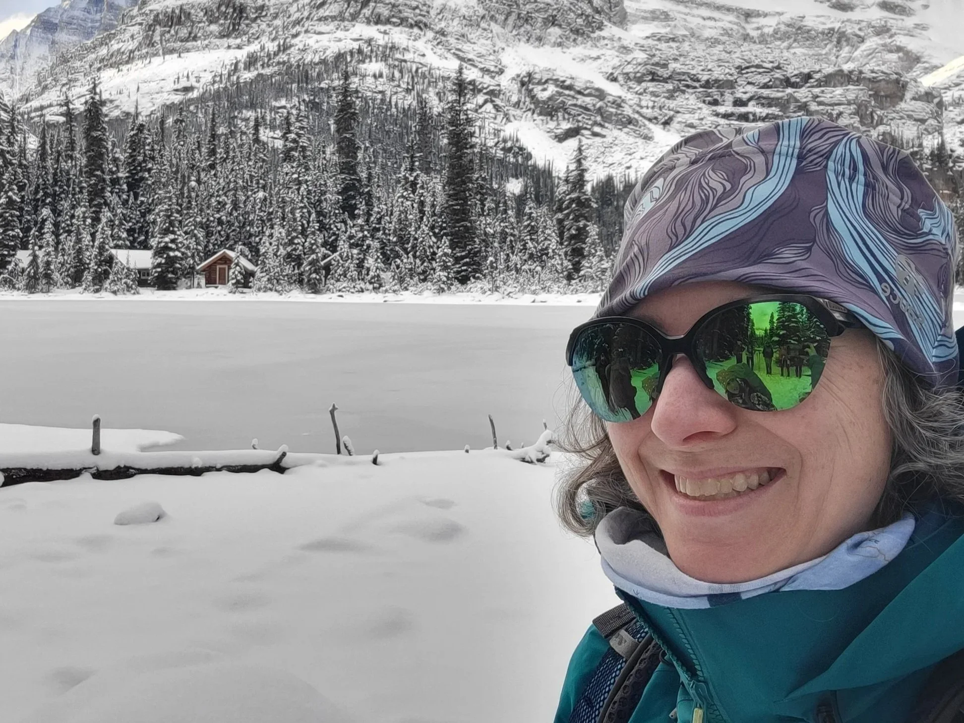 Woman in sunglasses and winter hat smiling, standing in snowy landscape with frozen lake, pine trees, and snow-covered mountains in background.