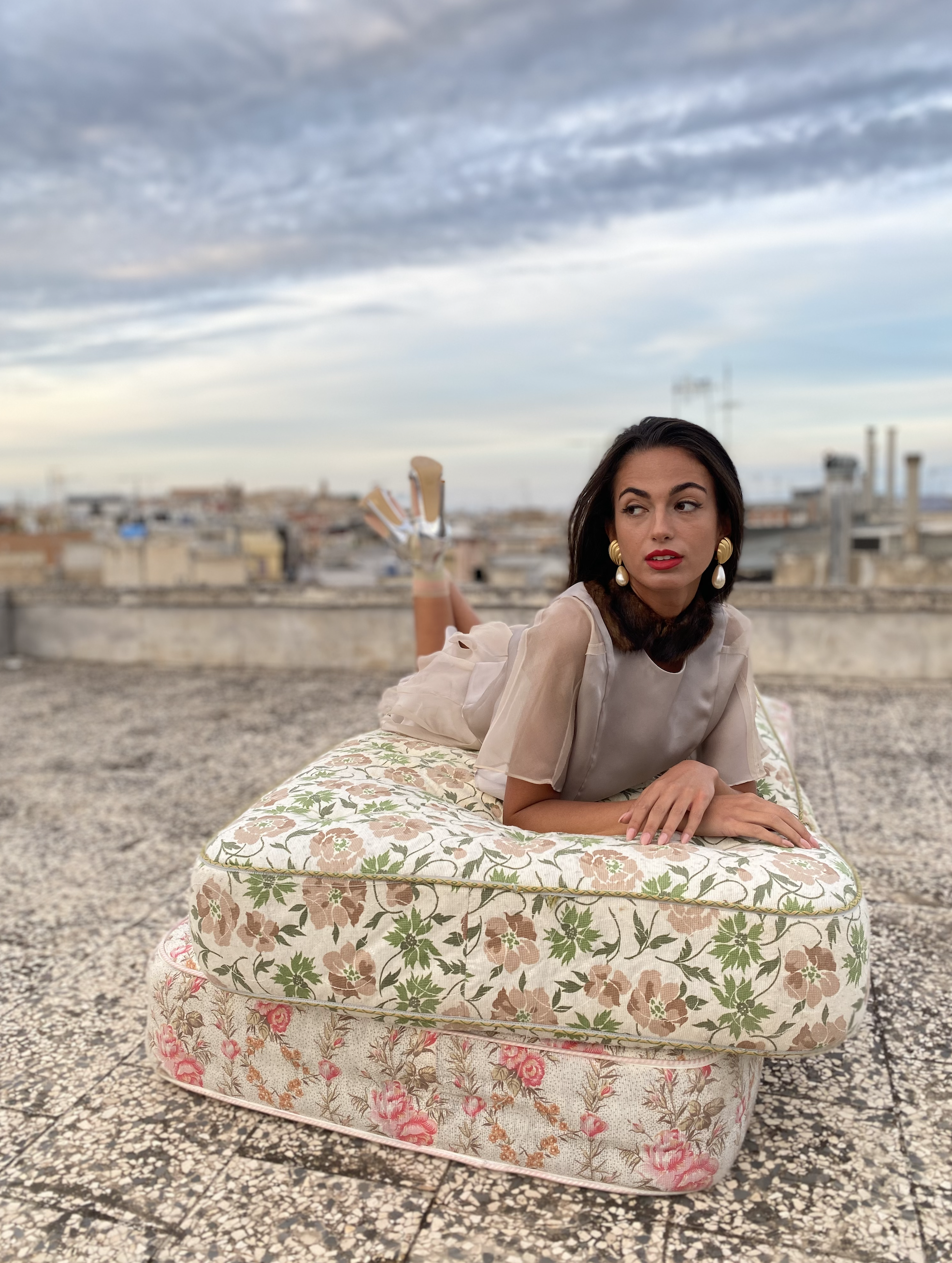 Model lying on a floral patterned mattress on a rooftop during sunset, with a cityscape and cloudy sky in the background. She is wearing large earrings, a light-colored Dreams in an Armoire Giuseppina silk organza top and Maria skirt.