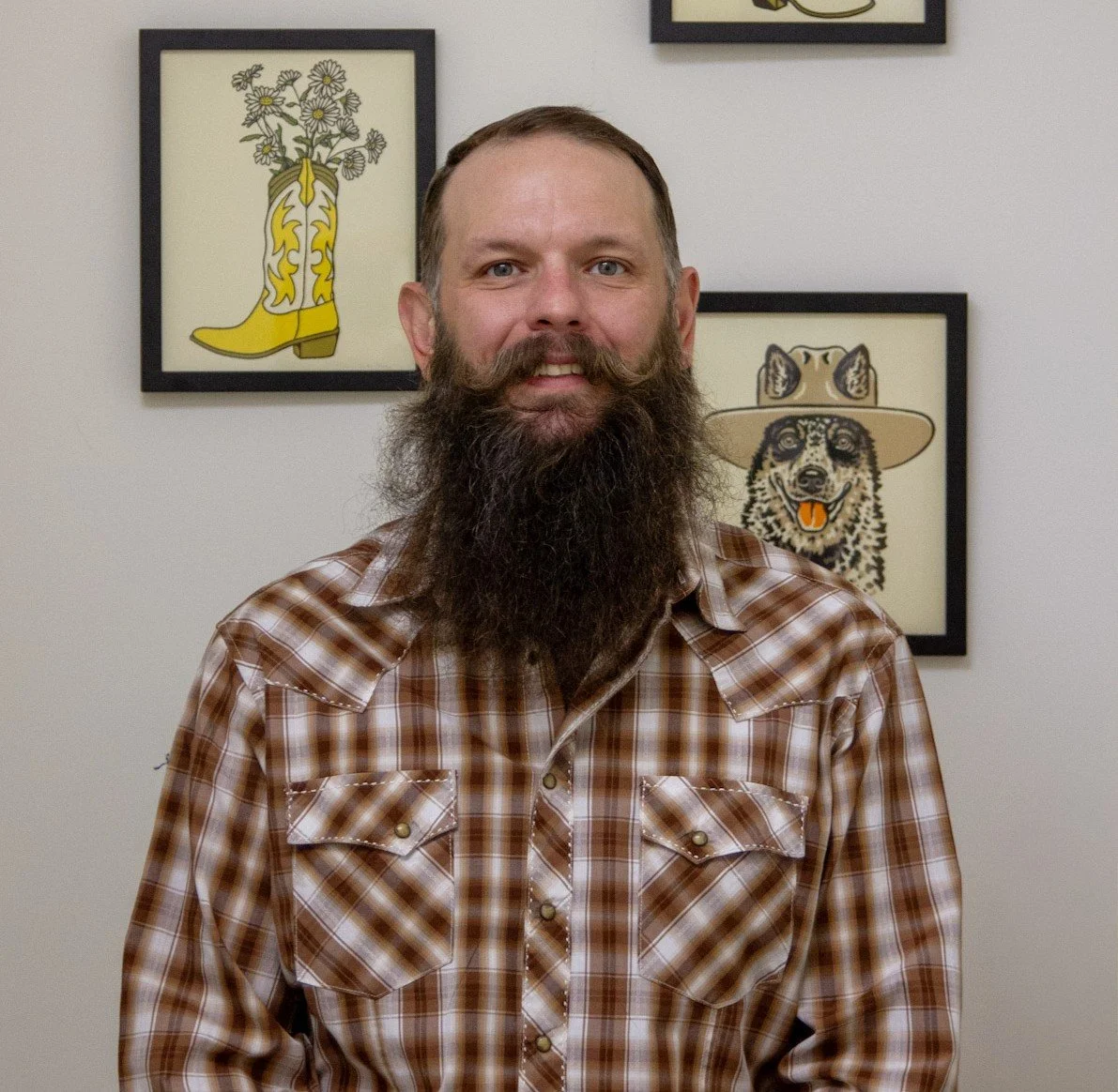 A man with a long beard and mustache wearing a plaid shirt standing in front of framed art depicting a yellow cowboy boot with flowers and a dog with a hat.