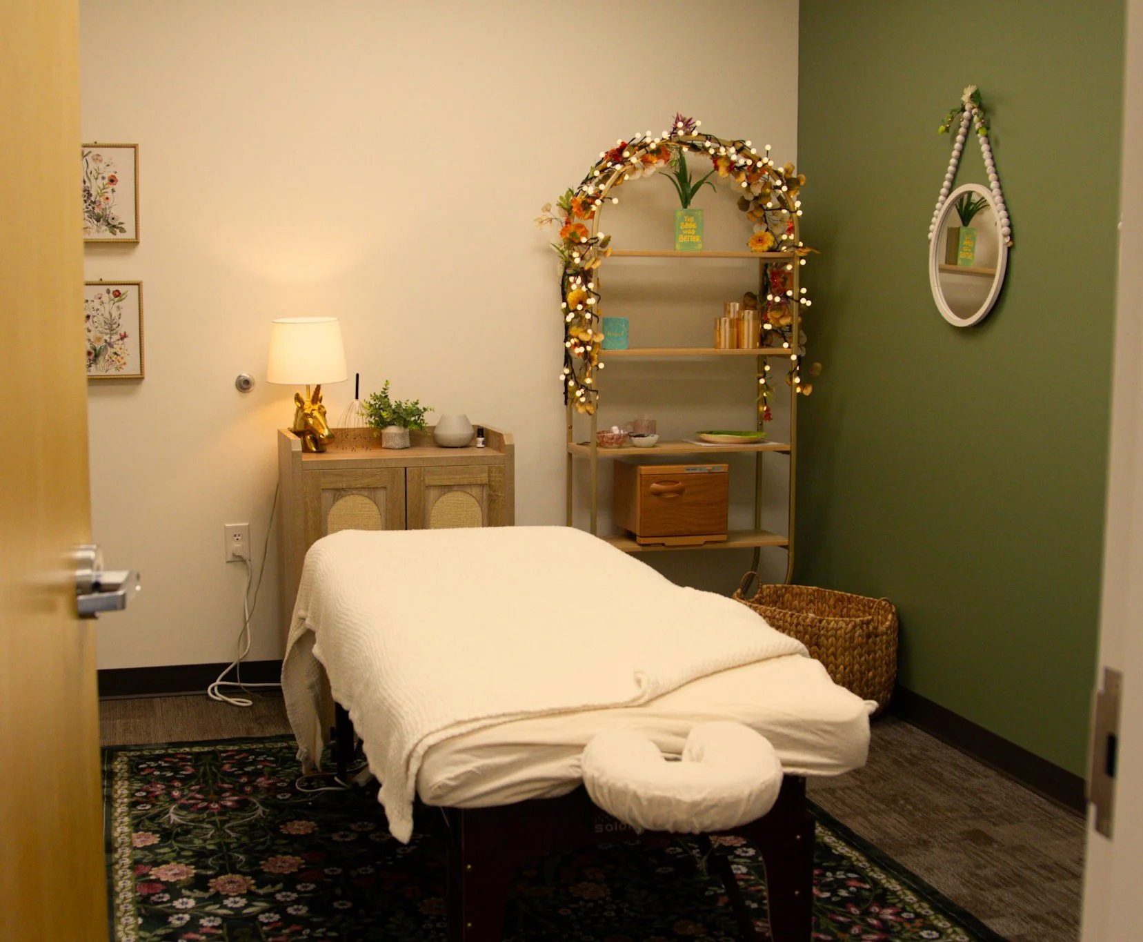 A cozy room with a massage table covered by a white blanket, a small ottoman at the foot, a patterned rug underneath, a wooden side table with a lamp and decor, a wicker basket beside the massage table, a decorative shelf with string lights, plants, and art on the walls.