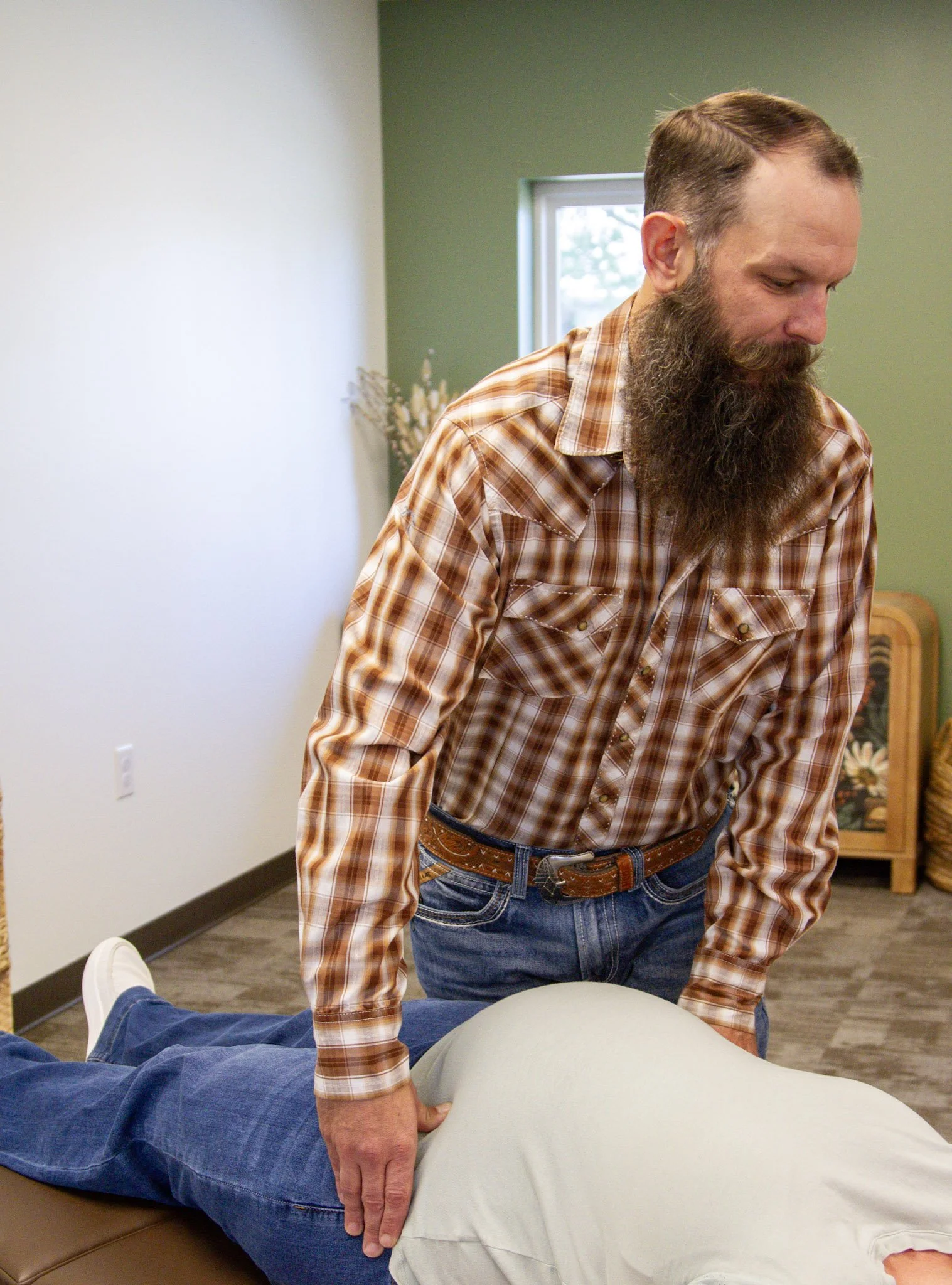 A man with a beard wearing a brown plaid shirt and jeans performs a chiropractic adjustment on a person's back who is lying face down on a table.