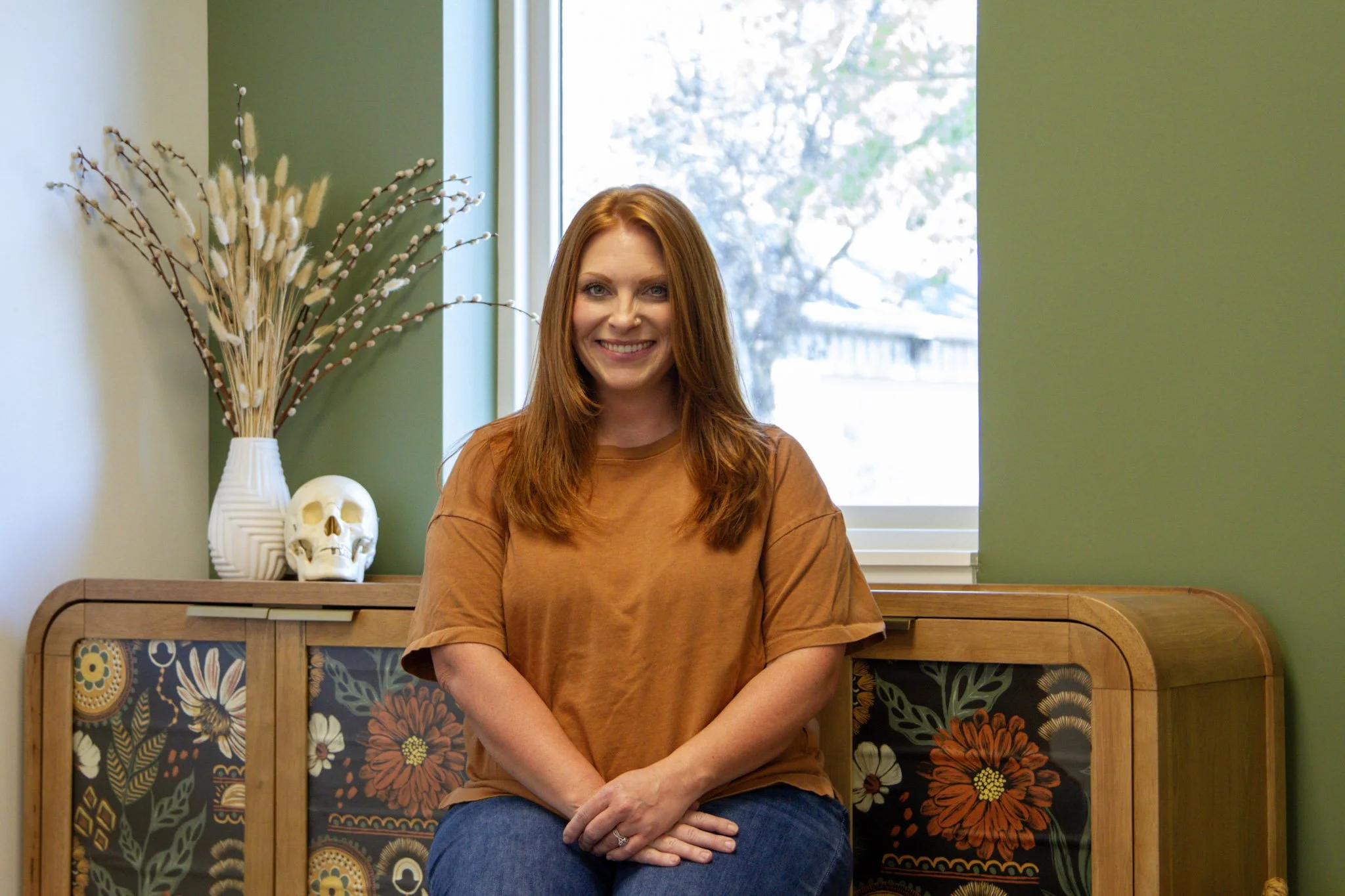 A woman with long red hair smiling, sitting on a colorful floral bench in front of a green wall with a white window behind her.