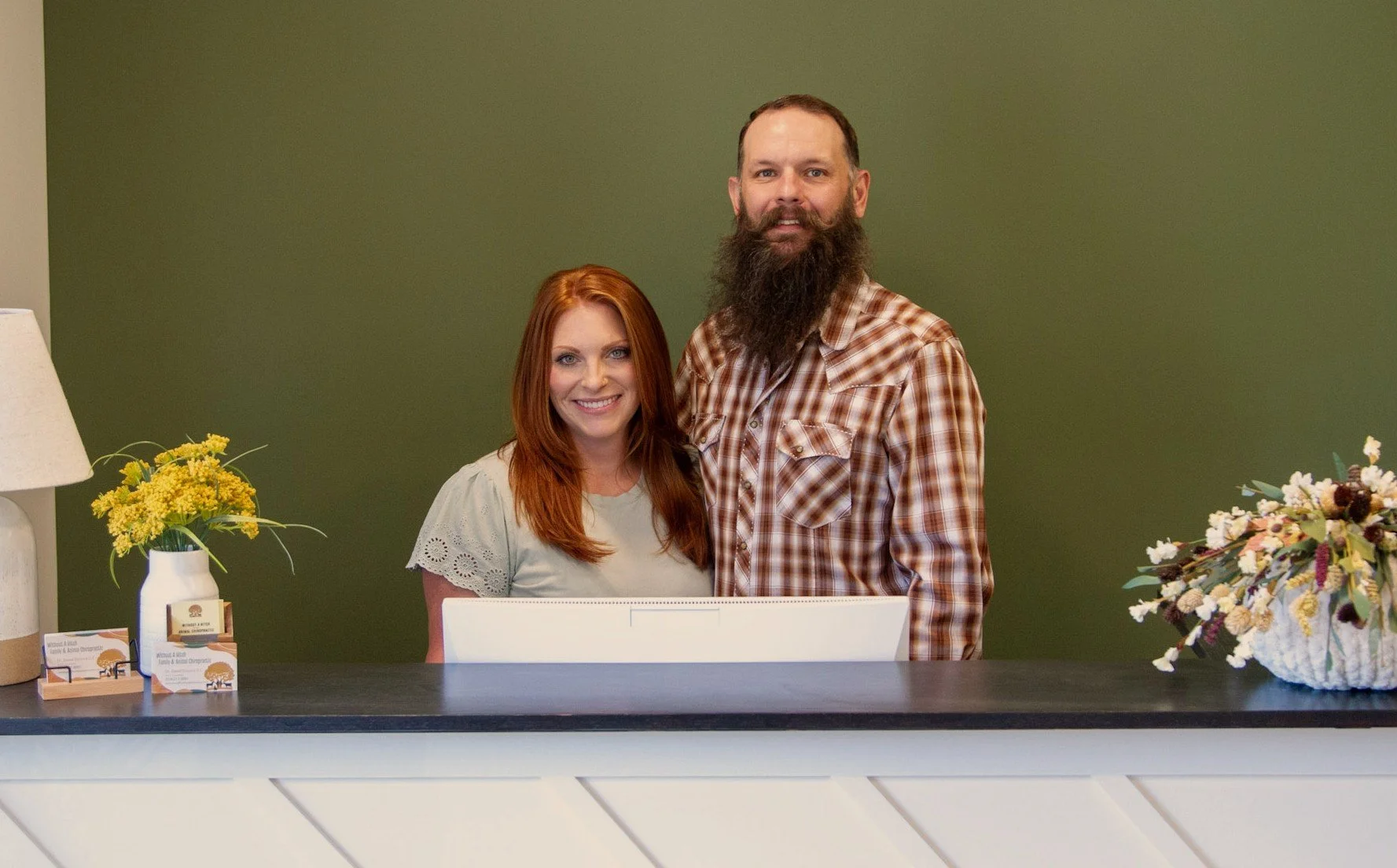 A smiling woman with long red hair and a man with a beard, standing behind a reception desk with a green wall in background. There are yellow flowers on the left and a mixed flower arrangement on the right.