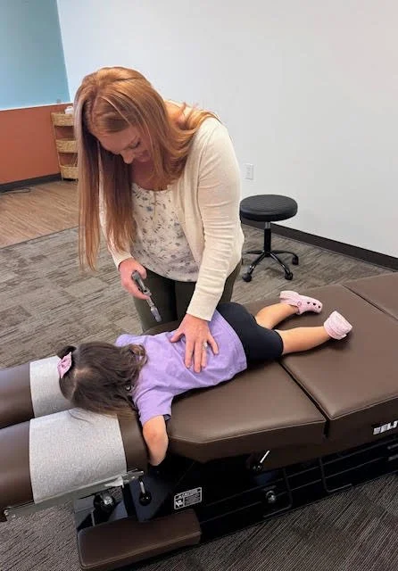 A woman is performing a medical or chiropractic adjustment on a young girl lying face down on a treatment table, in a clinical setting.