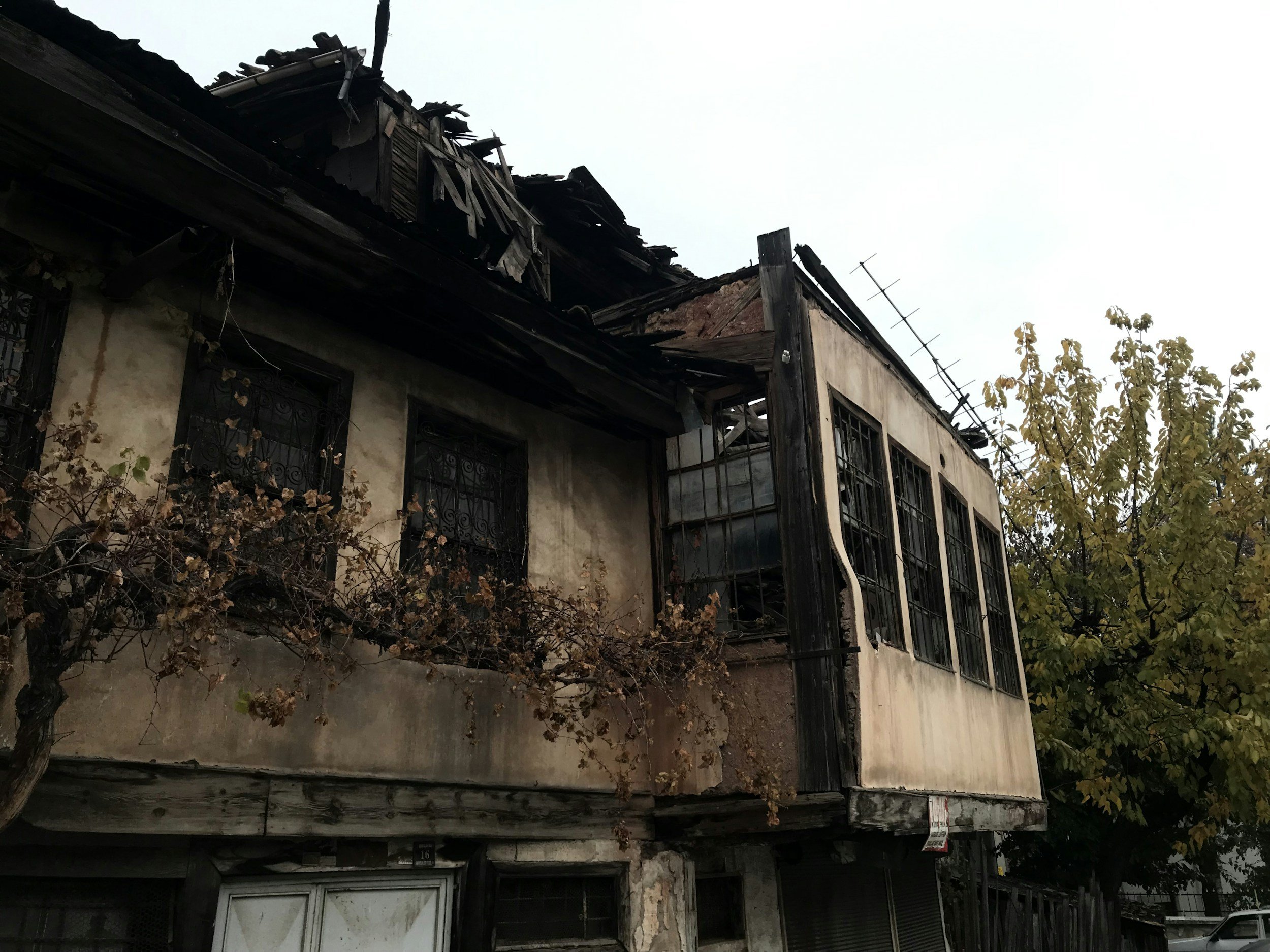 A partially burned two-story house with charred exterior and broken windows, with a tree in front and a gray sky above.