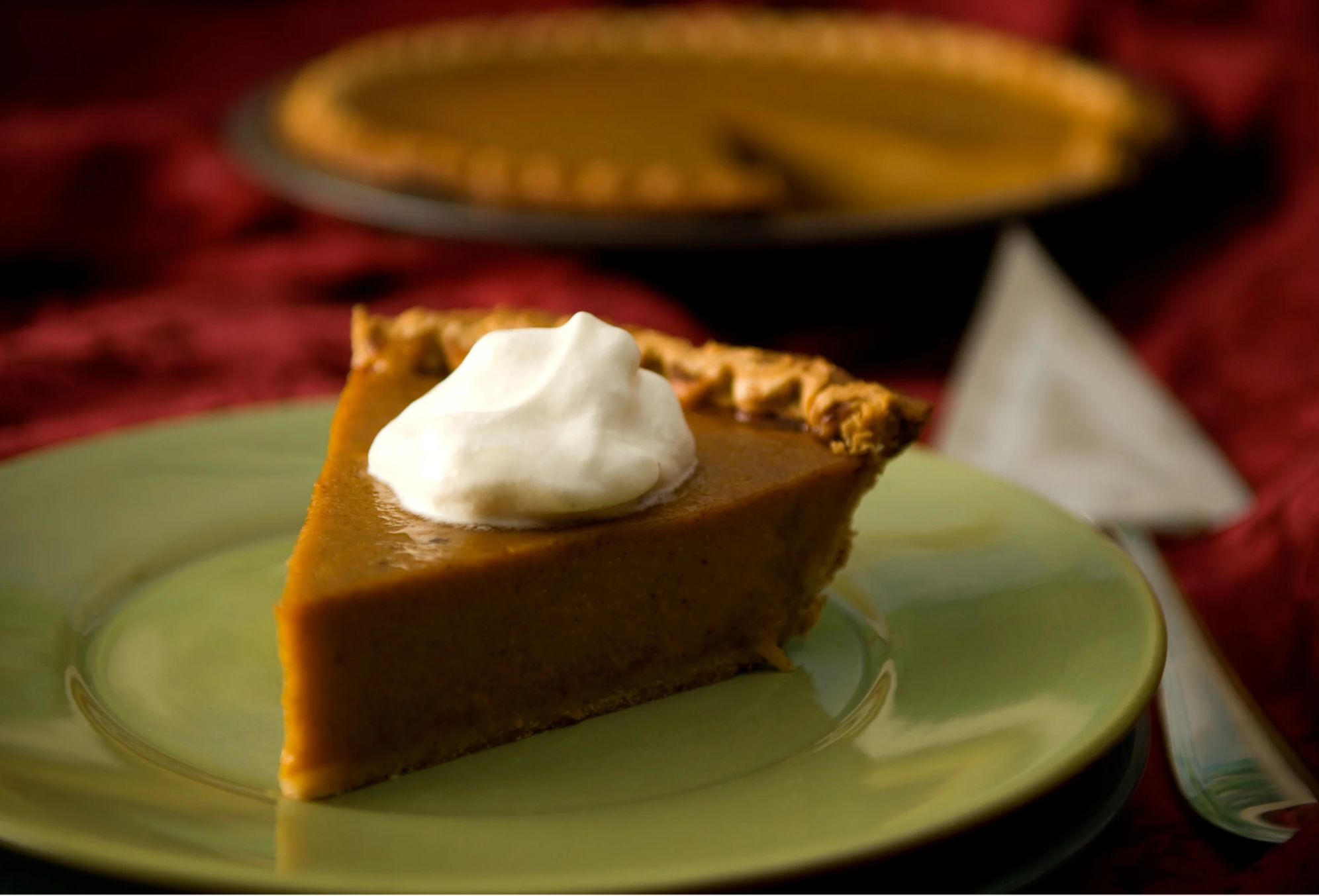 A slice of pumpkin pie topped with whipped cream on a green plate, with the whole pie in a black dish in the background.