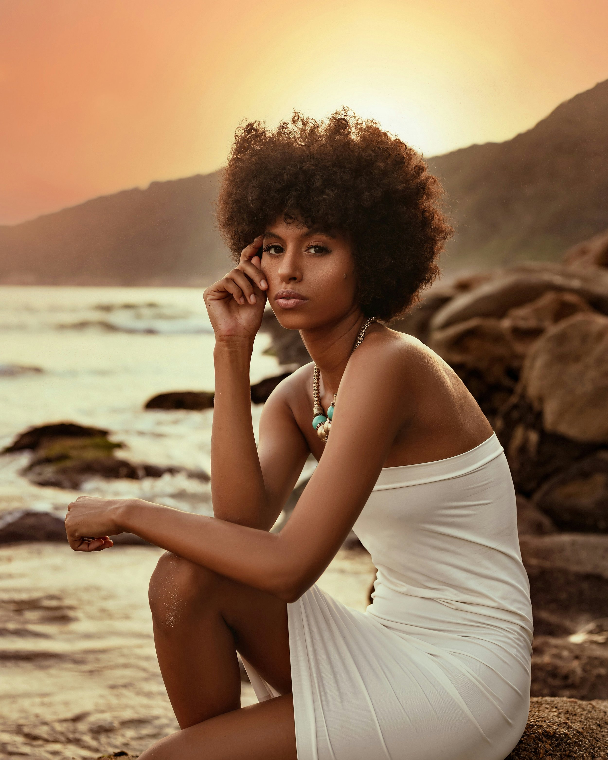 A woman with curly hair sitting on rocks on the beach at sunset, wearing a white strapless dress and jewelry.
