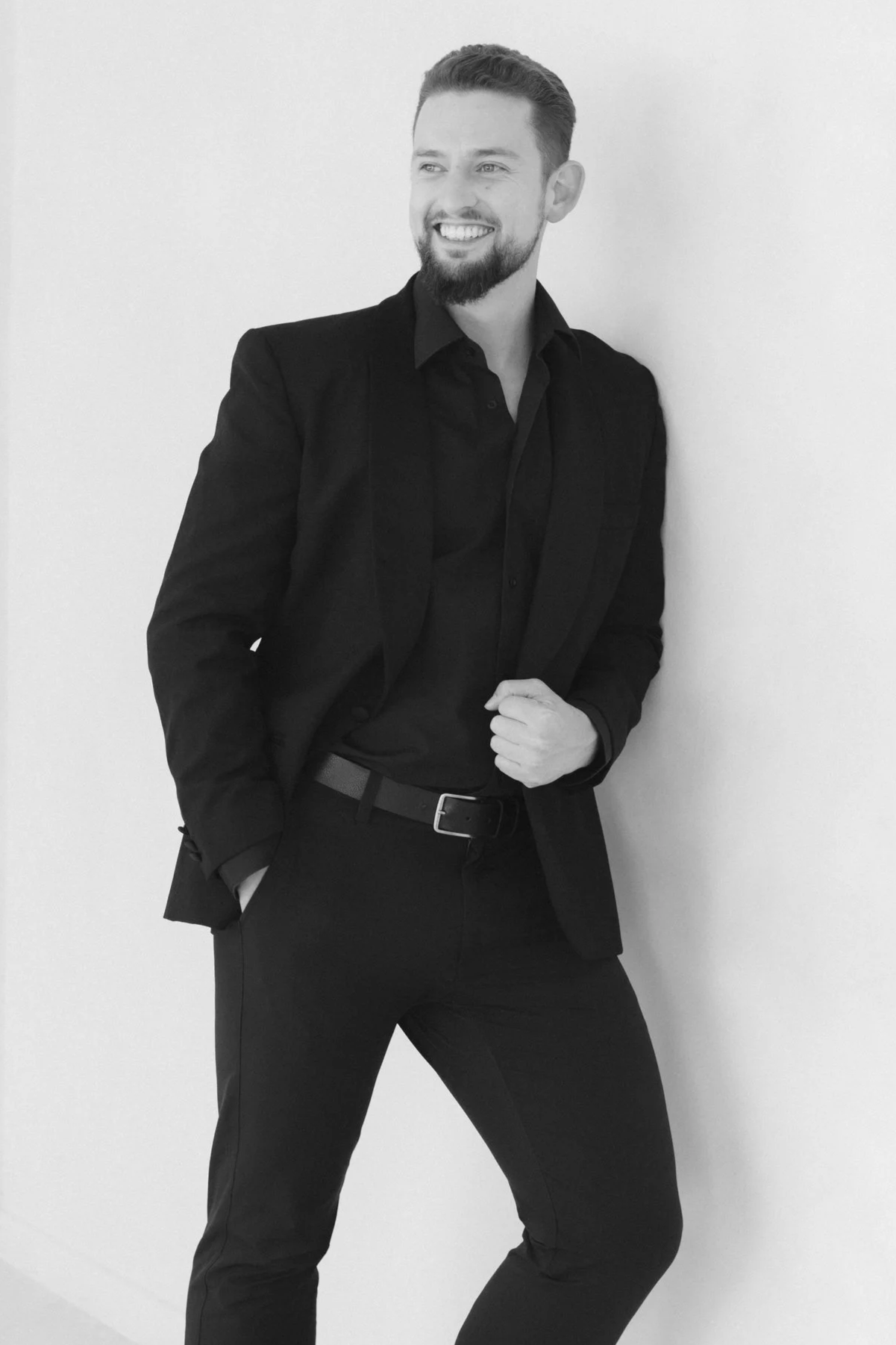 Clements In Motion posing with a smile, displaying a genuine confident, happy expression in a smart black suit in a studio setting. The picture is in black and white.