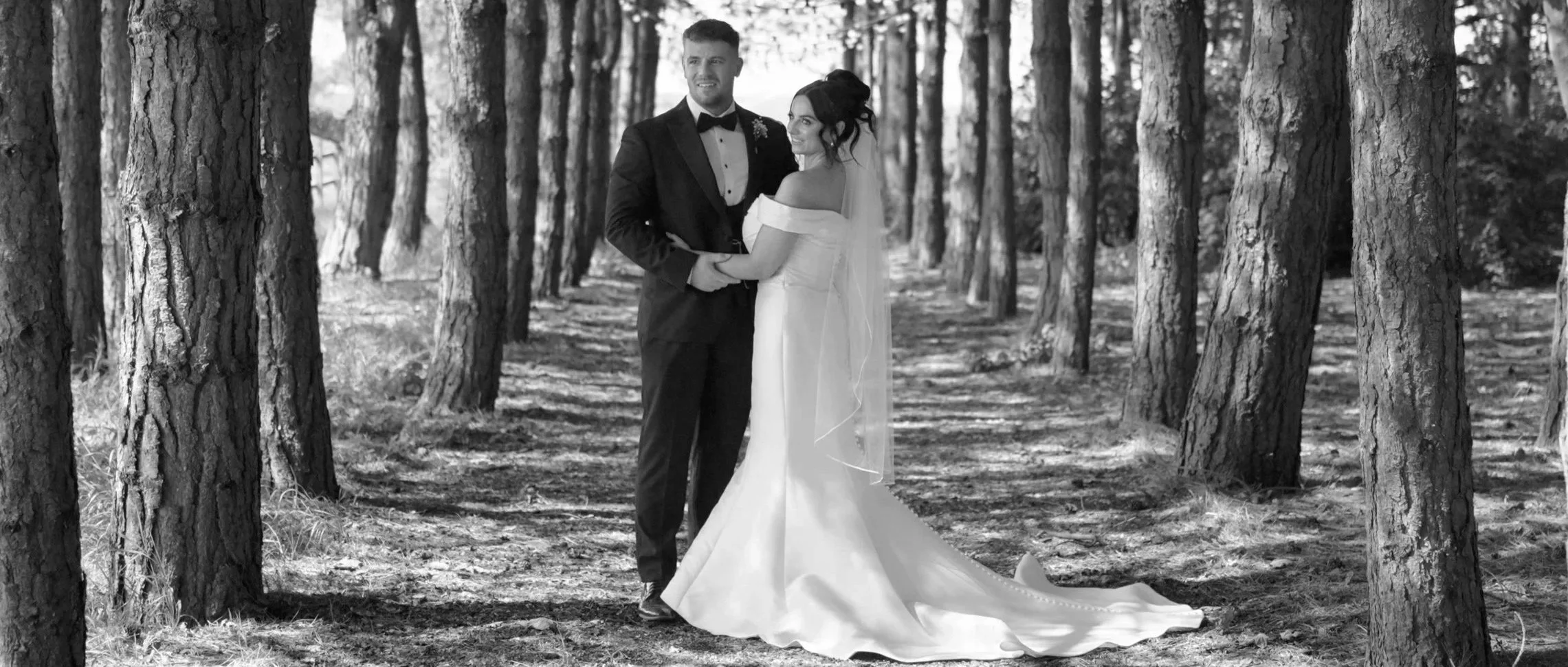 The Kynes, a bride and groom standing together in a wooded area at Bunny Hill Weddings, the groom wearing a tuxedo and the bride in a wedding dress, holding hands and smiling.