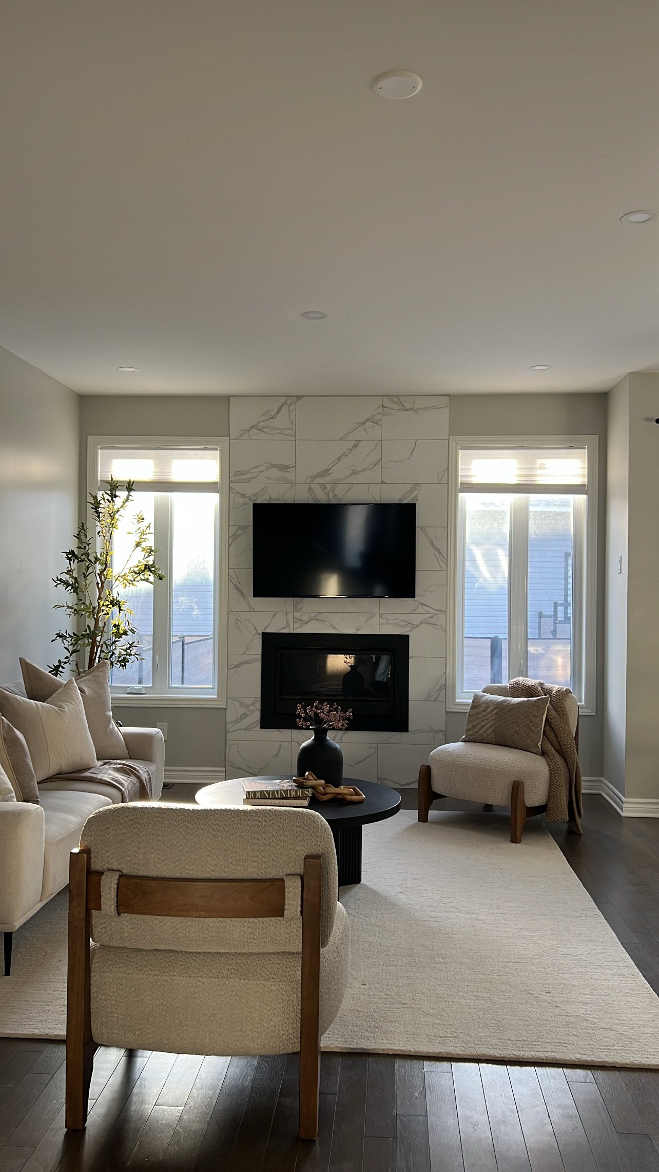 Living room with a fireplace, wall-mounted TV, two large windows, beige sofa, armchair, black coffee table with a vase and books, and a cream rug on dark hardwood floor.