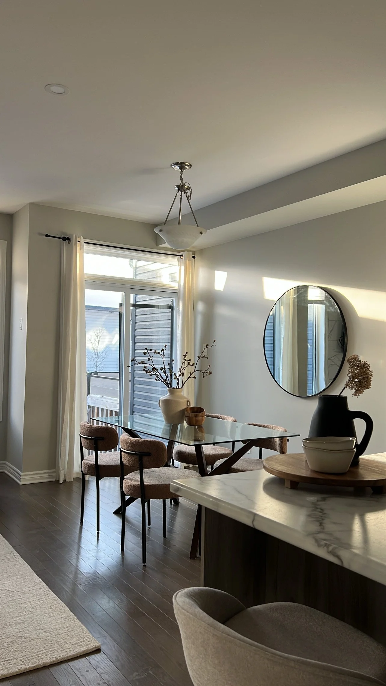 Interior view of a dining area with a glass table, pink upholstered chairs, a round mirror on the wall, a vase with branches, and a sliding glass door with curtains allowing sunlight into the room.