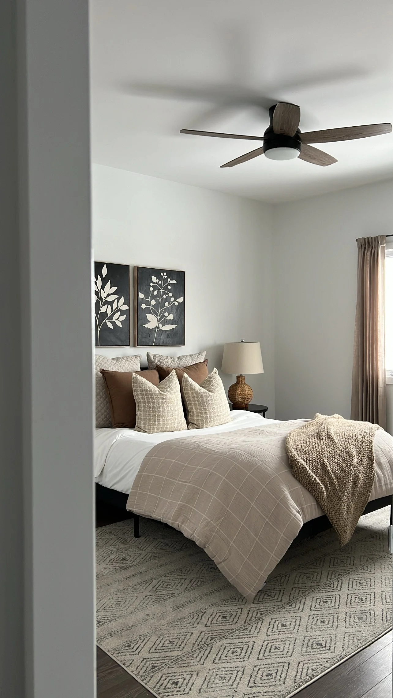 A bedroom with a bed, multiple beige and brown pillows, a beige blanket, a nightstand with a woven lamp, wall art of two black-framed floral prints, a window with beige curtains, a ceiling fan, and a patterned area rug.