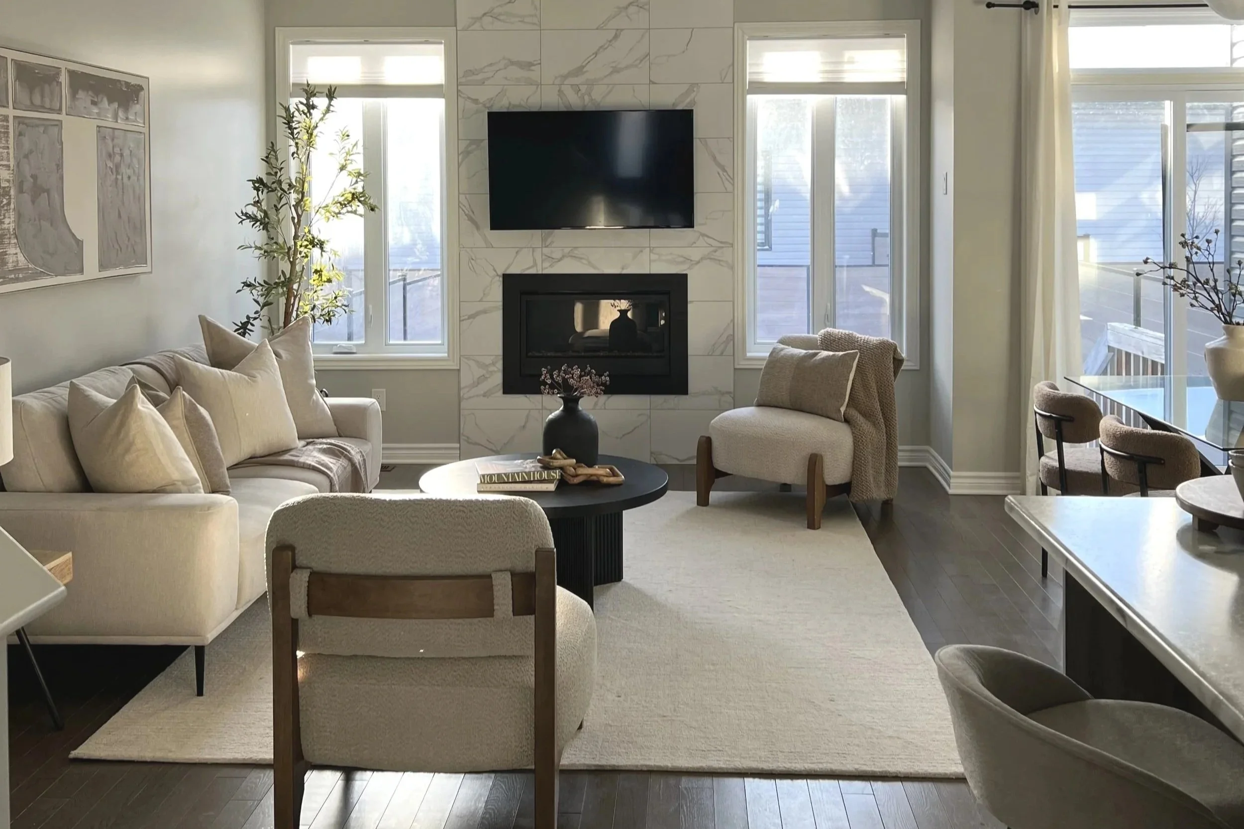 Living room with cream-colored sofa, beige armchair, black coffee table, and a fireplace with a mounted TV above. Large windows on either side brightening the space, with sliding glass door on the right. Potted plants and artwork decorate the room, a