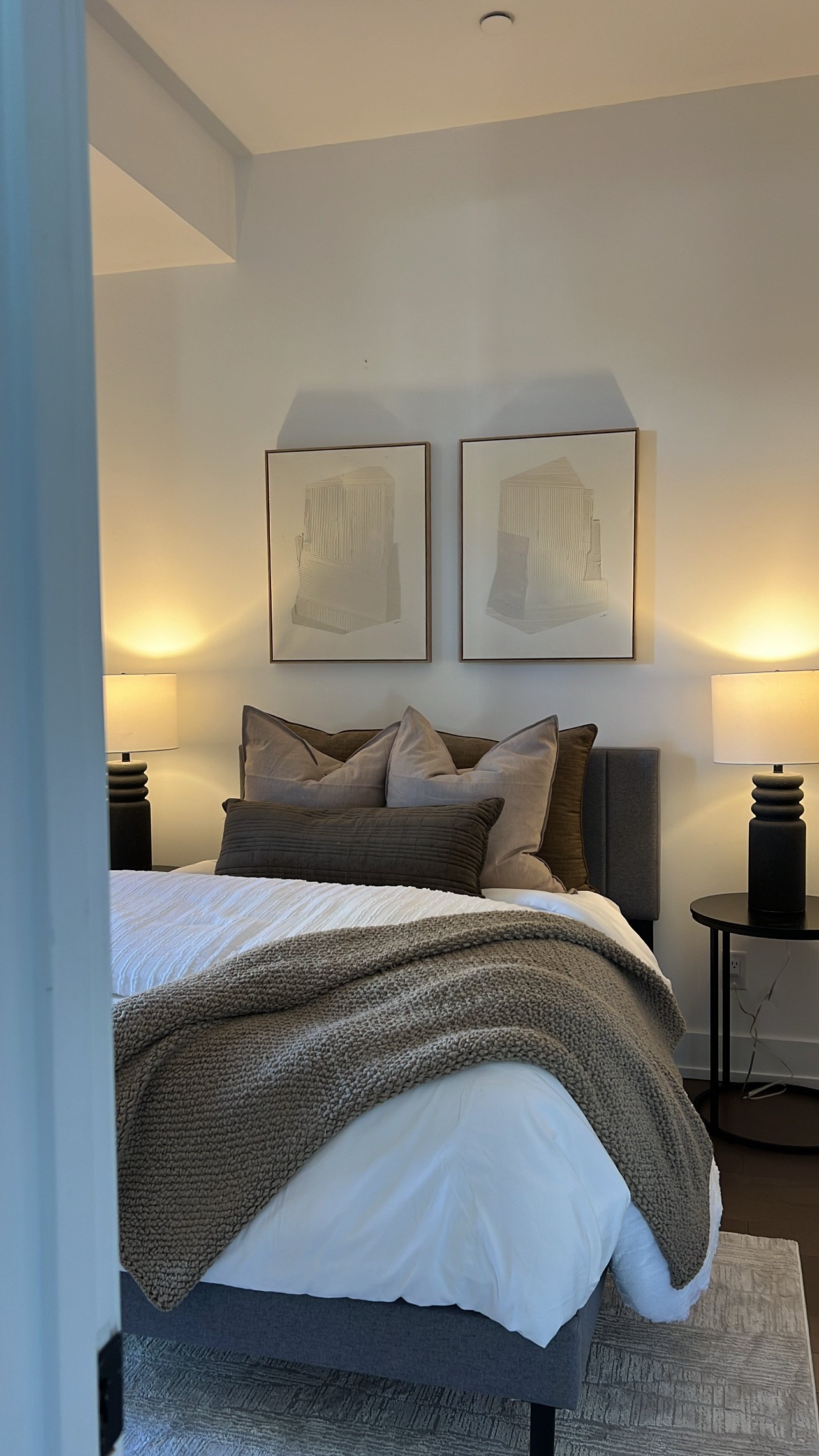 A cozy bedroom with a bed, decorative pillows, a knitted throw blanket, two nightstands with lamps, and framed artwork on the wall.