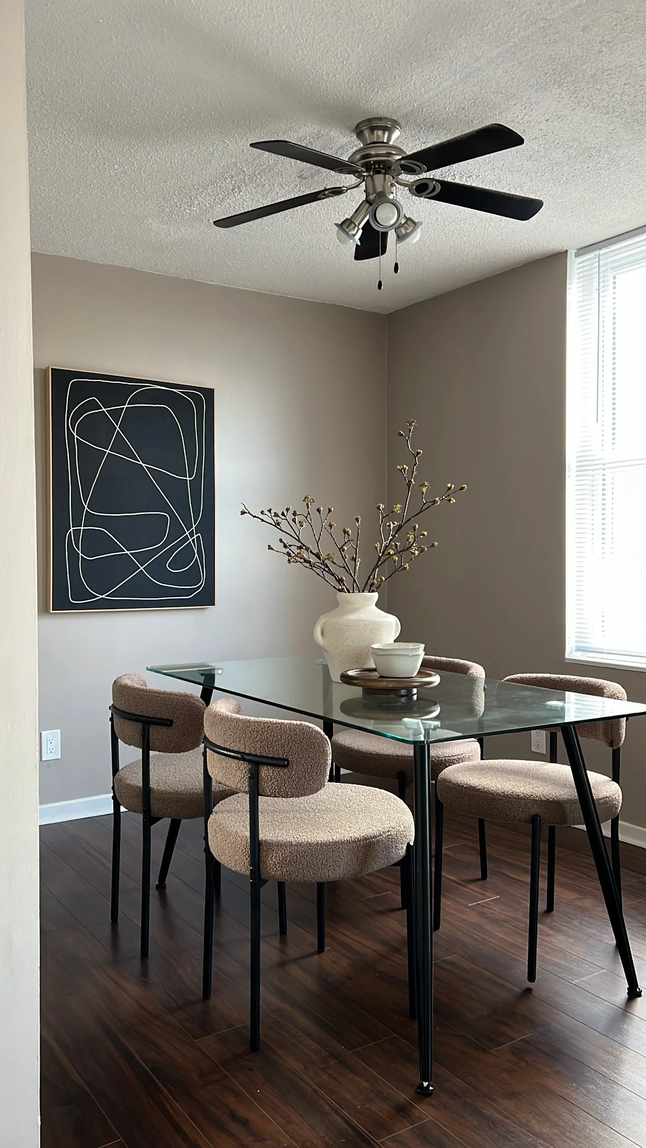 A dining room with a glass-top table, six cushioned chairs, a large white vase with branches, a black and white abstract wall art, a ceiling fan, and a window with blinds.