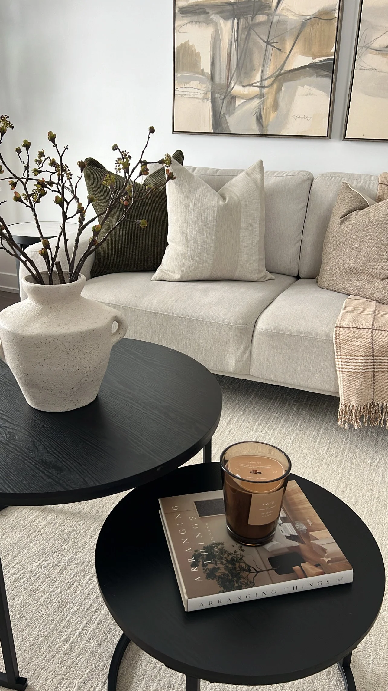Living room with a beige sofa, decorative pillows, abstract wall art, black coffee tables, a book titled 'Arranging Things', a candle, and a ceramic vase with branches.