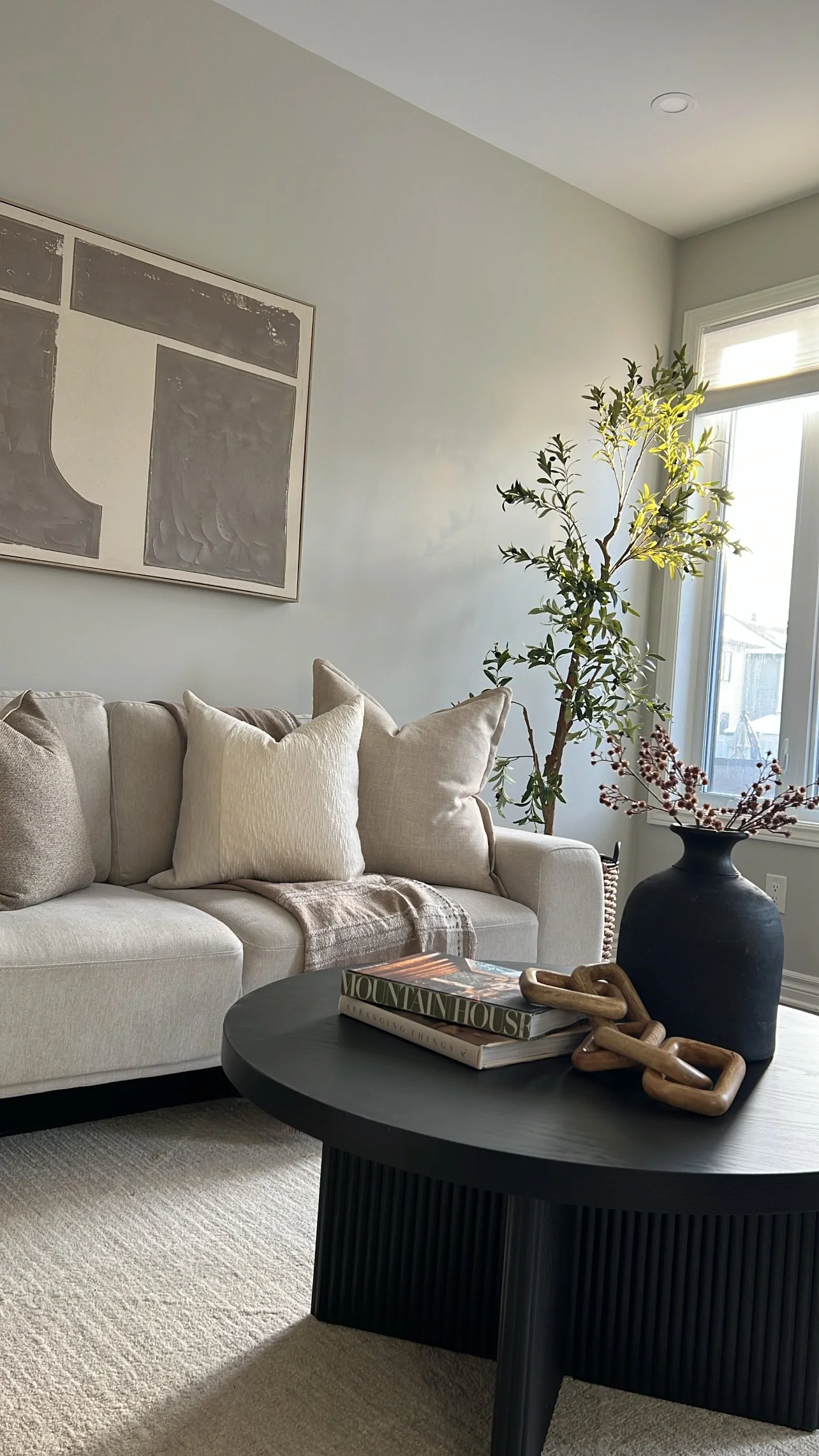 Living room with beige sofa, beige pillows, black coffee table with books, large black vase with pink flowers, tall plant near window, light-colored walls, abstract wall art, sunlight coming through window.