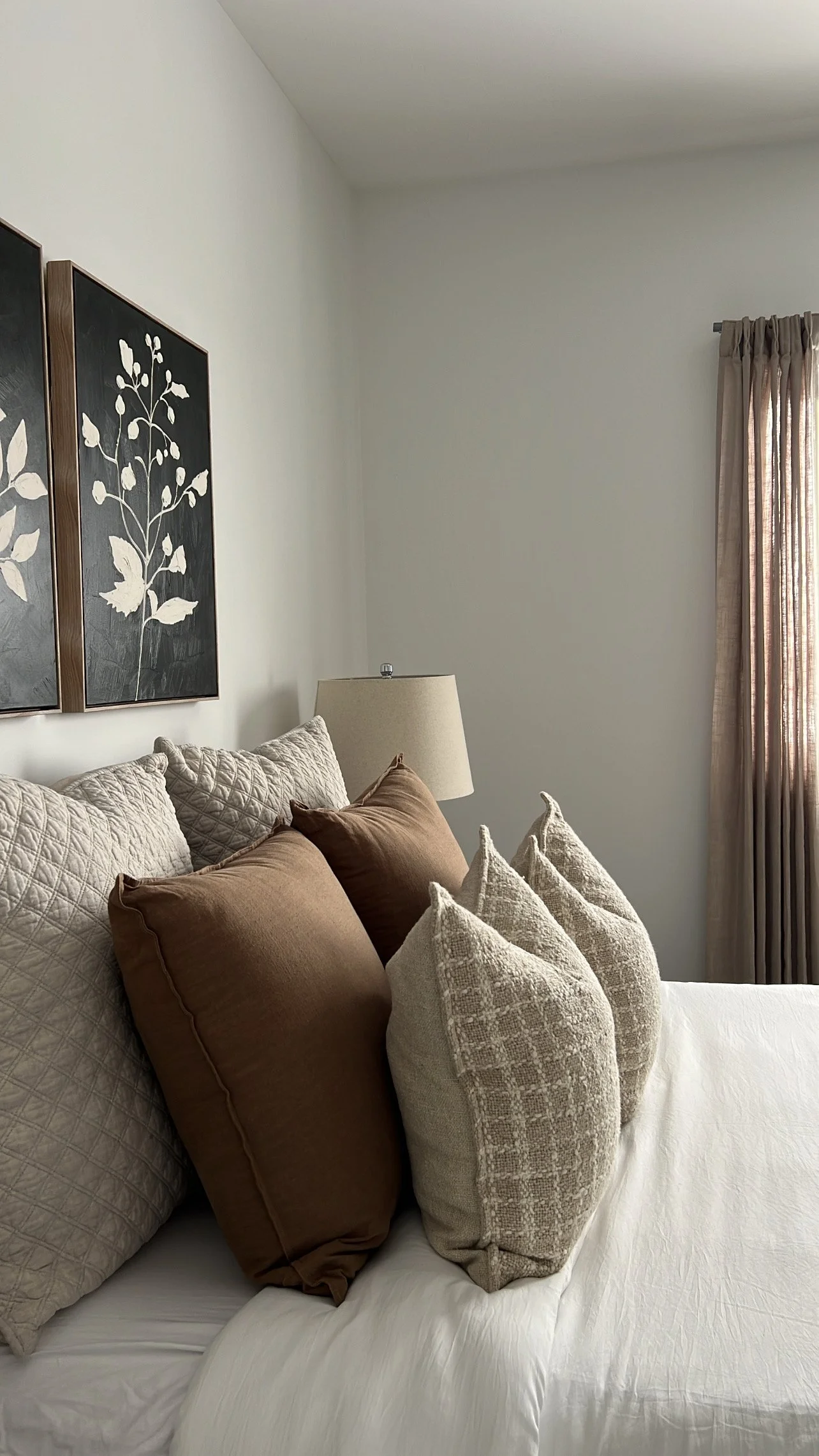 A neatly made bed with brown and beige pillows, a bedside lamp, wall art featuring botanical designs, and a window with beige curtains.
